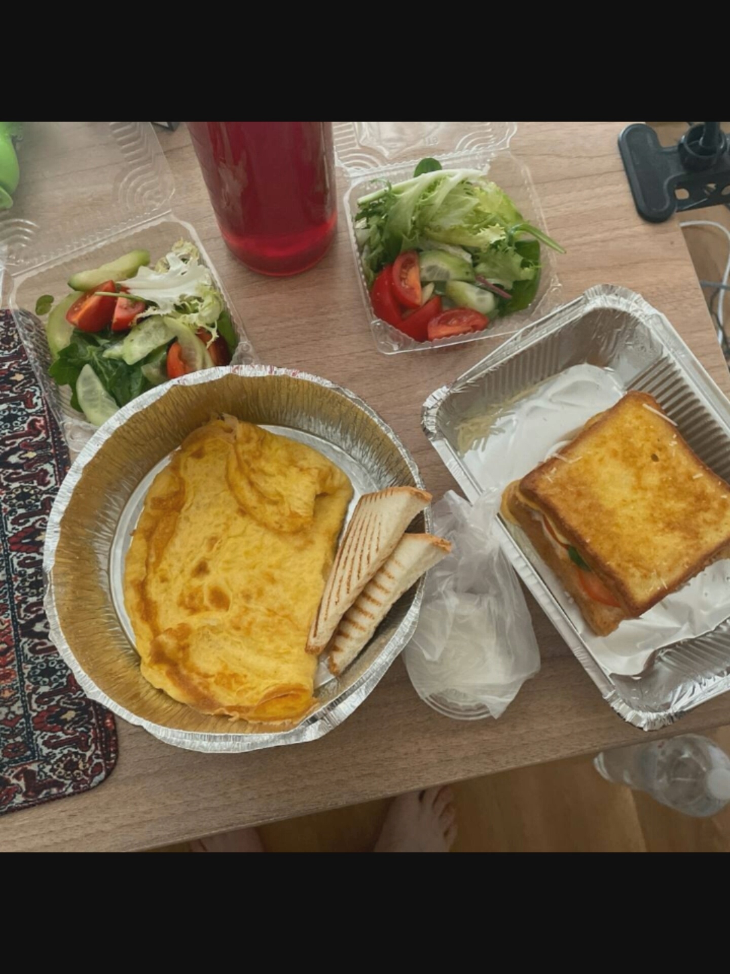 A table is laden with a meal, suggesting a casual dining experience. The scene is set on a wooden surface, likely a table, with hints of a patterned rug or cloth on the left. The lighting suggests daytime.
The subjects are primarily food items arranged in various containers. In the foreground, a round aluminum foil container holds a golden-brown omelet, partially folded, alongside several triangular pieces of grilled toast. To the right of the omelet, another aluminum foil container presents a grilled sandwich, golden brown and appearing to have cheese and tomato inside. Adjacent to these main dishes, two clear plastic containers hold salads. The salad in the left container is a mix of lettuce, cucumber slices, and cherry tomatoes. The salad in the right container is similar, with lettuce and tomatoes visible. A tall glass containing a deep red liquid, possibly a juice or beverage, stands behind the salads. A small, clear plastic bag filled with a white, granular substance, possibly sugar or salt, sits between the omelet and the grilled sandwich. A partially visible clear plastic bottle with a white cap lies on the wooden surface towards the bottom right.
The activity appears to be a meal being prepared for or consumed. The presence of takeout containers and the arrangement on the table suggest either a takeaway meal or a casual home-cooked meal served in a practical manner. The overall impression is one of a simple, satisfying meal. No people are clearly visible, though a bare foot is peeking into the frame at the bottom. There is no visible text in the image. The context provided mentions Bucharest, Romania, but no specific cues within the image strongly suggest this location.