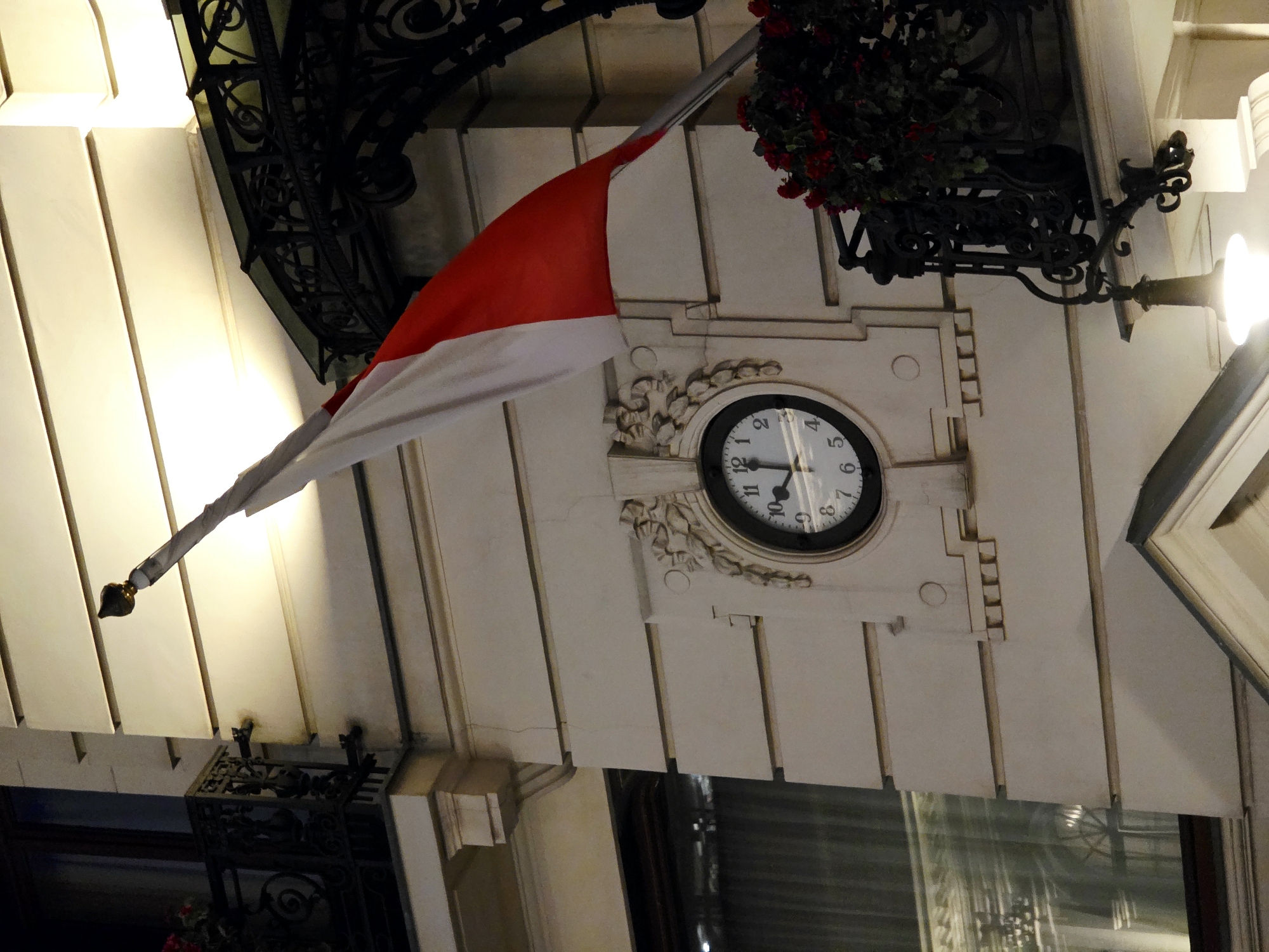 The image shows a close-up, exterior view of a building's facade at night in Warsaw, Poland. The building is made of light-colored stone with horizontal banding and decorative elements. A white and red flag is partially visible, extending from the top left and waving to the right. Below the flag, a circular clock is embedded in the wall, showing the time as approximately 10:09. The clock is surrounded by an ornate, sculpted frame. To the right of the clock, there is a black wrought-iron balcony with a planter overflowing with red flowers. A decorative black metal awning is visible above the balcony. The lighting in the scene is warm and artificial, suggesting streetlights or building illumination. The overall impression is one of an elegant, historic building.