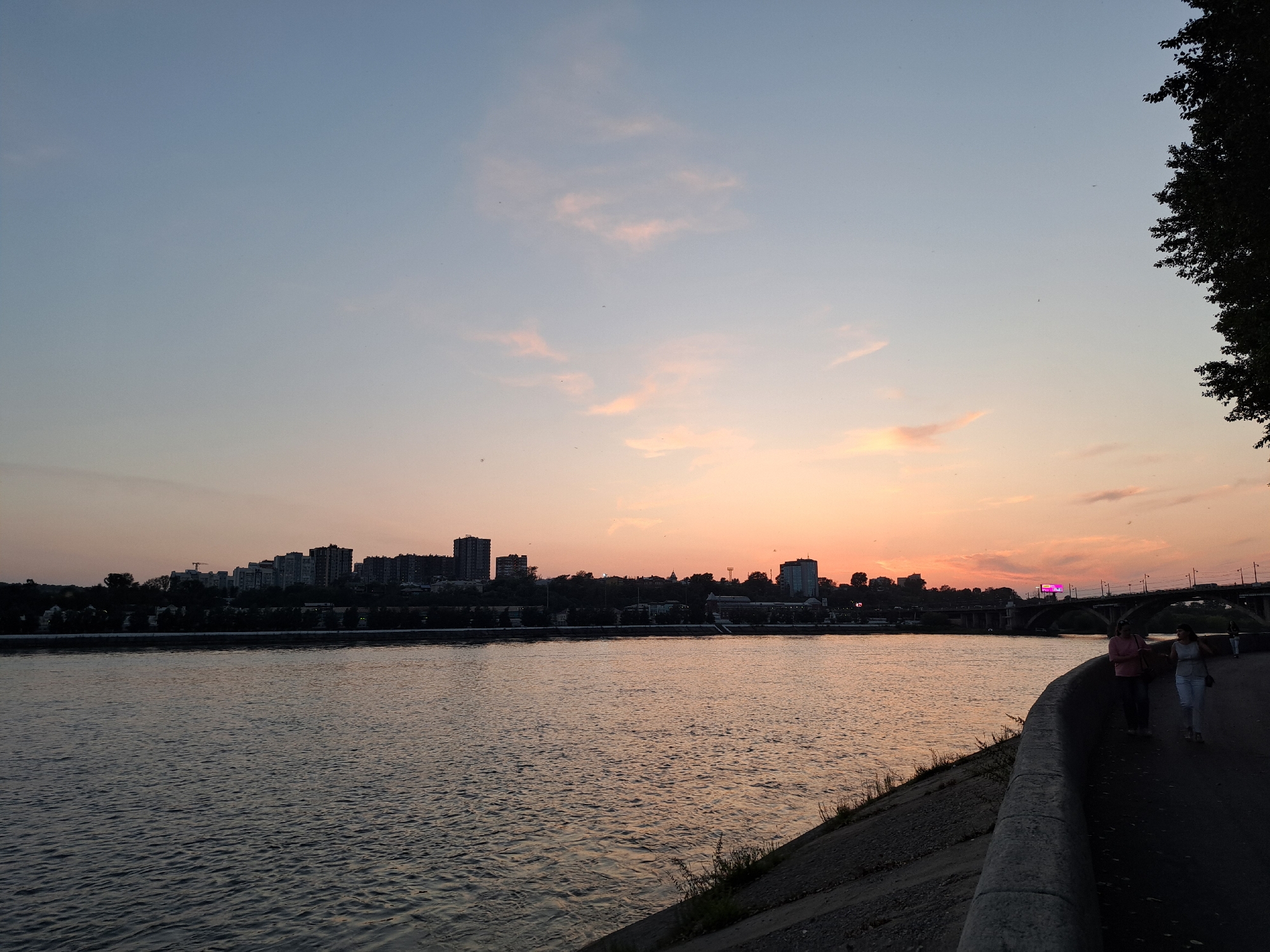 A photograph captures a sunset over the Angara River in Irkutsk, Russia. Silhouetted against the warm hues of the setting sun, a cityscape with numerous buildings and apartment blocks lines the opposite bank. A bridge spans the river in the distance. In the foreground, two women walk along a paved embankment, their figures cast in shadow. The river's surface reflects the soft light of the sky, with gentle ripples creating a textured pattern. The scene is set against a backdrop of a gradient sky, transitioning from pale blue at the top to warm oranges and pinks near the horizon.
