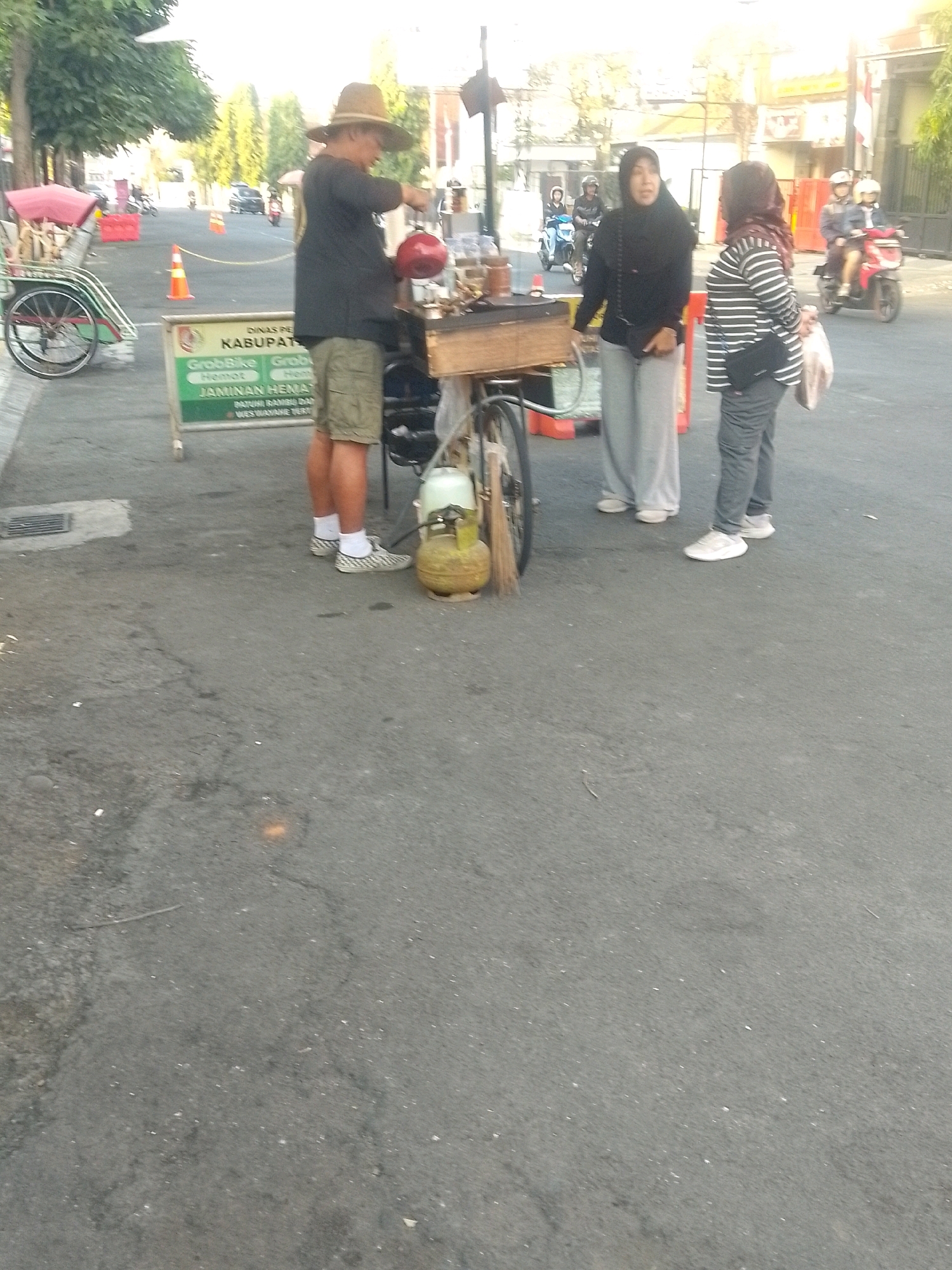 The image shows a street scene in Jember, Indonesia. In the foreground, a man wearing a straw hat, a dark shirt, and khaki shorts stands by a food cart. He is holding a red container. To his right is a gas cylinder and a broom. Behind the man, a woman in a black hijab and light gray pants stands next to another woman wearing a striped long-sleeved shirt and dark pants. Both women are looking towards the man. The food cart has a sign on it with text that is partially visible. Further in the background, a green and red pedicab is parked on the left. On the right side of the street, several people are on motorcycles, and trees line the background. The pavement is asphalt, and there is a slight incline to the street. The lighting suggests it is daytime, likely late afternoon.