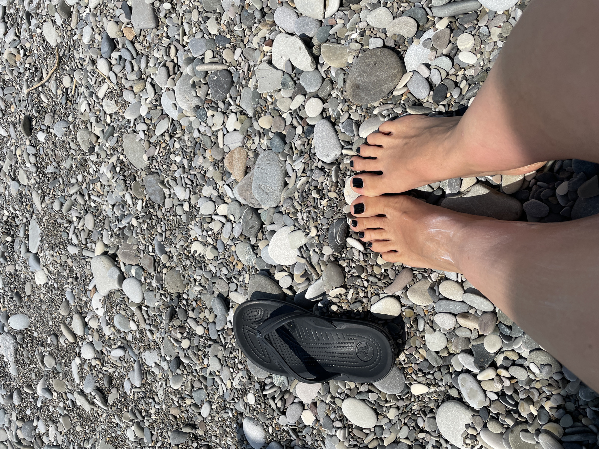 A top-down shot of a person's feet on a pebbly beach, with a black flip-flop next to them. The person's toenails are painted black. The beach is covered in various sizes and colors of smooth, round pebbles. The lighting suggests it is a sunny day. The location is unknown.