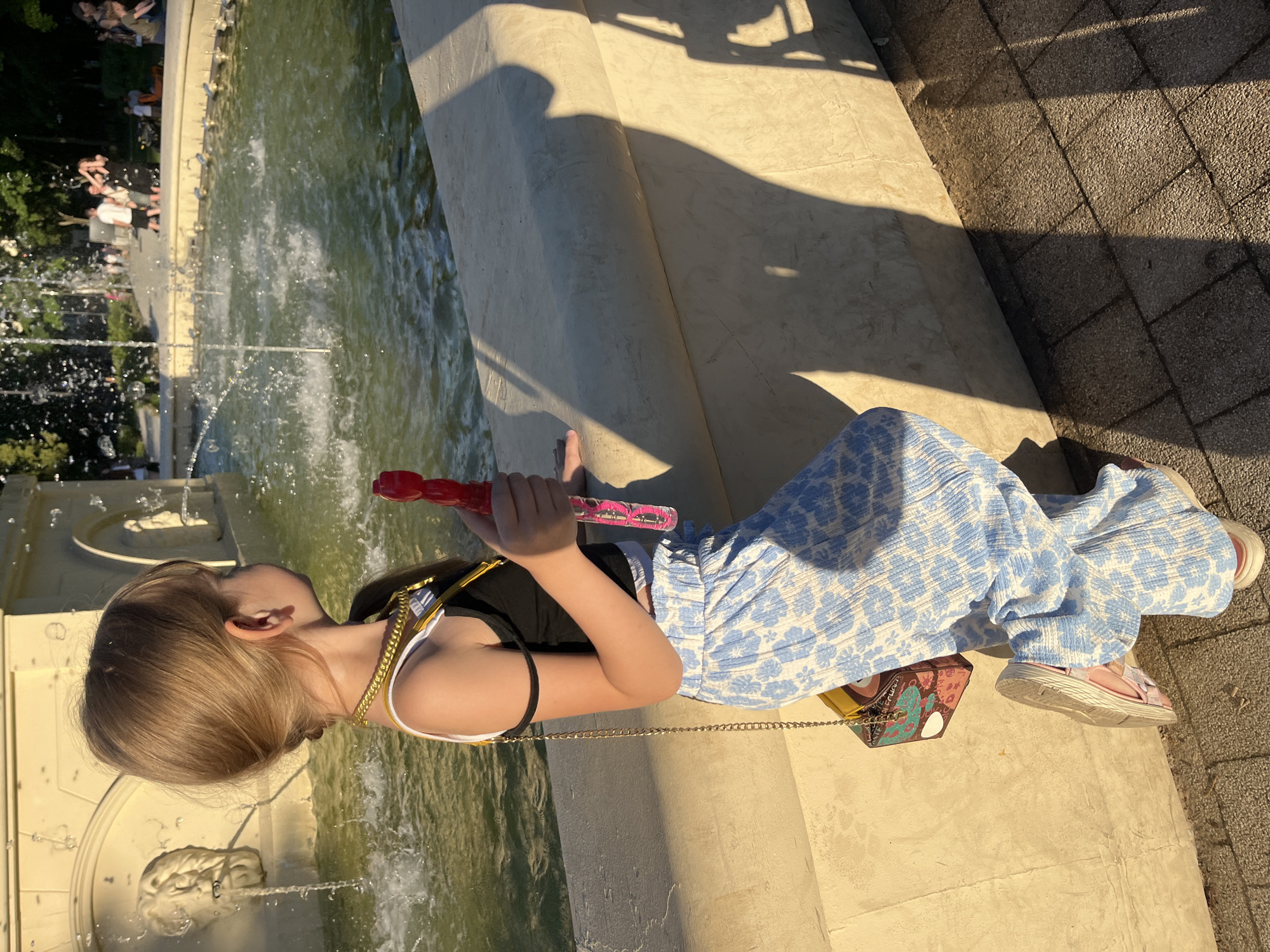 In Novaya Usman', Russia, a young girl with blonde hair pulled into a ponytail stands by a fountain. She is wearing a black tank top with a white undershirt, a gold chain necklace, and light blue pants with a floral pattern. In her right hand, she holds a red and pink bubble gun. A small, colorful purse with a gold chain strap hangs from her left shoulder. Her left foot is propped up on a stone ledge, and her right foot is on the ground. The fountain has water spraying, and in the background, there are trees and people walking. The scene is bathed in warm sunlight, casting shadows on the ground.