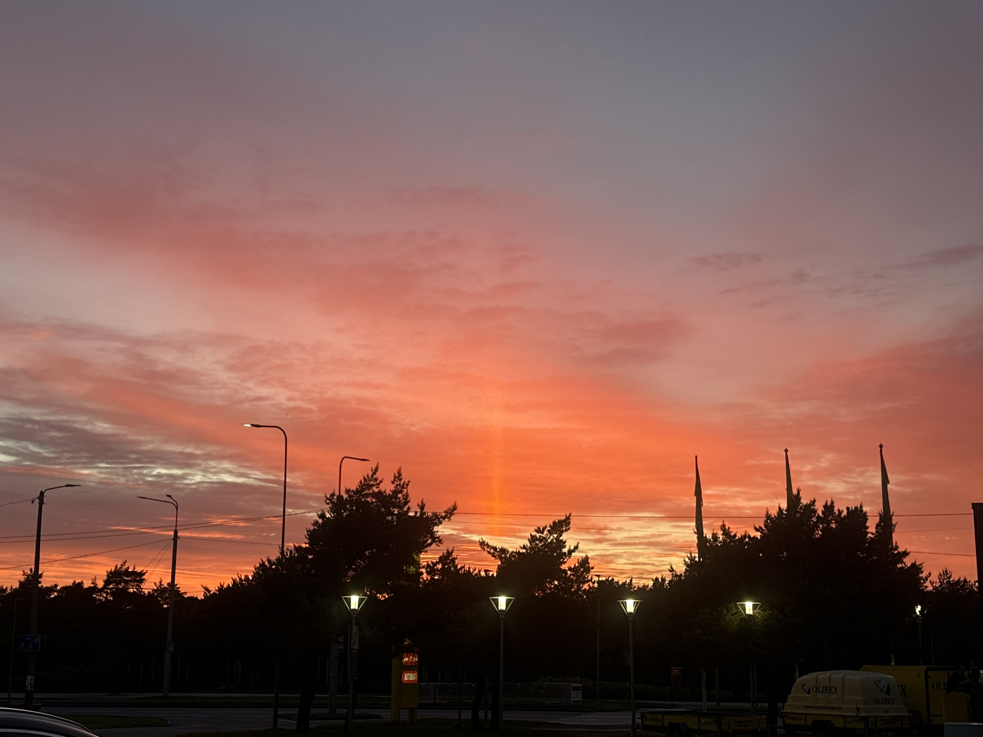 A vibrant sunset paints the sky in hues of orange, pink, and purple over a treeline in Tallinn, Estonia. Silhouetted against the colorful sky are several streetlights and tall trees, some with flags planted amongst them. In the foreground, there are glimpses of a road, a parking area, and a yellow trailer with the text "OLIREX" printed on it. The scene appears to be captured in an outdoor urban or suburban setting, possibly near a business complex given the trailer and flags.