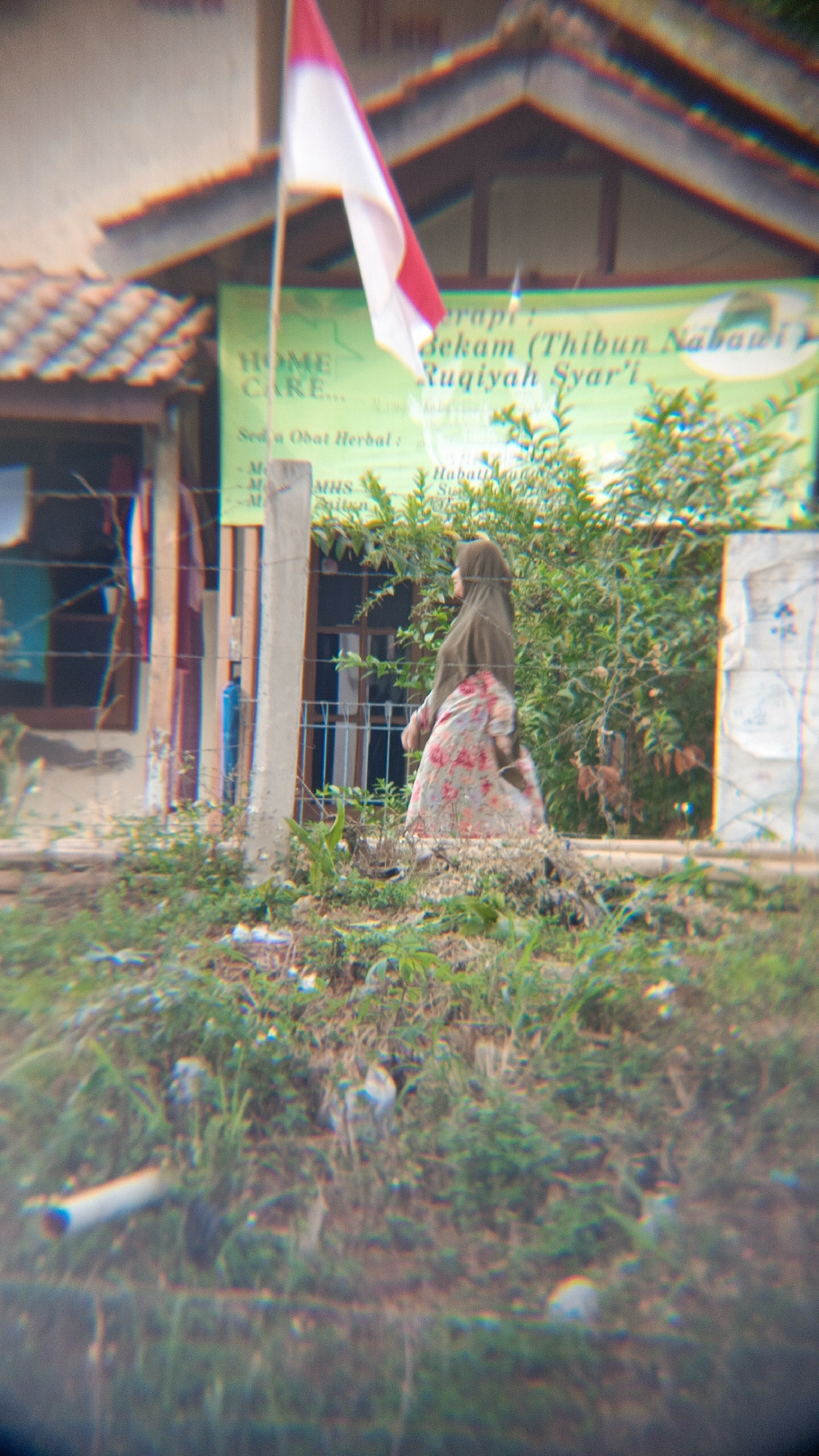In the city of Margajaya, Indonesia, an outdoor scene unfolds with a focus on a person in Islamic attire. The individual, a woman, is seen from behind, wearing a brown hijab and a floral patterned dress with pink and white flowers. She is walking away from the viewer and towards a lush green bush. A red and white Indonesian flag is prominently displayed on a pole, fluttering in the foreground, slightly out of focus. Behind the flag, a building with a tiled roof is visible, with a banner hanging from it that appears to contain text in Indonesian. The ground is covered in grass and other low-lying vegetation. A wire fence runs across the lower left quadrant of the image. The overall impression is one of a local street scene, possibly near a community center or place of business, with a strong cultural presence indicated by the flag and the woman's attire.