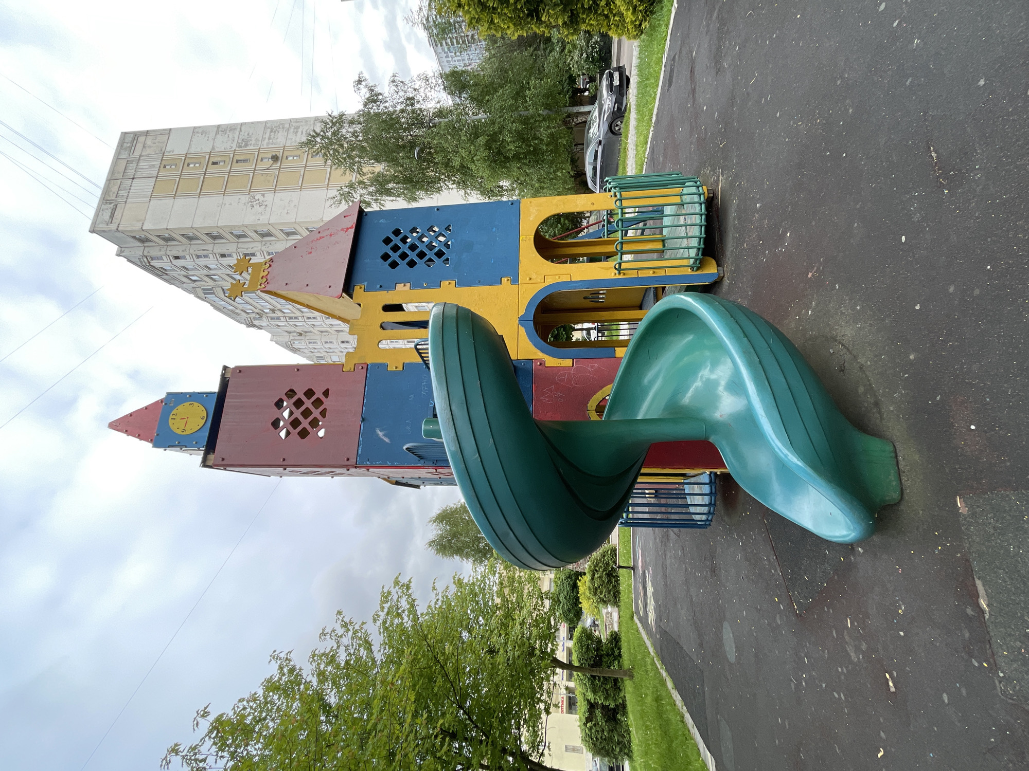 The image depicts a colorful playground structure with a winding slide in a park setting, likely in Moscow, Russia. The structure is composed of interconnected blocks in red, blue, and yellow, featuring geometric cutouts and a clock tower element. The slide is green and spirals downward. The playground is situated on a black asphalt surface with some visible wear and tear. In the background, there's a tall, light-colored building, partially obscured by trees. A few other trees and patches of greenery are visible, along with a blurred car parked near the edge of the park. The sky is overcast. No people are visible in the immediate foreground.