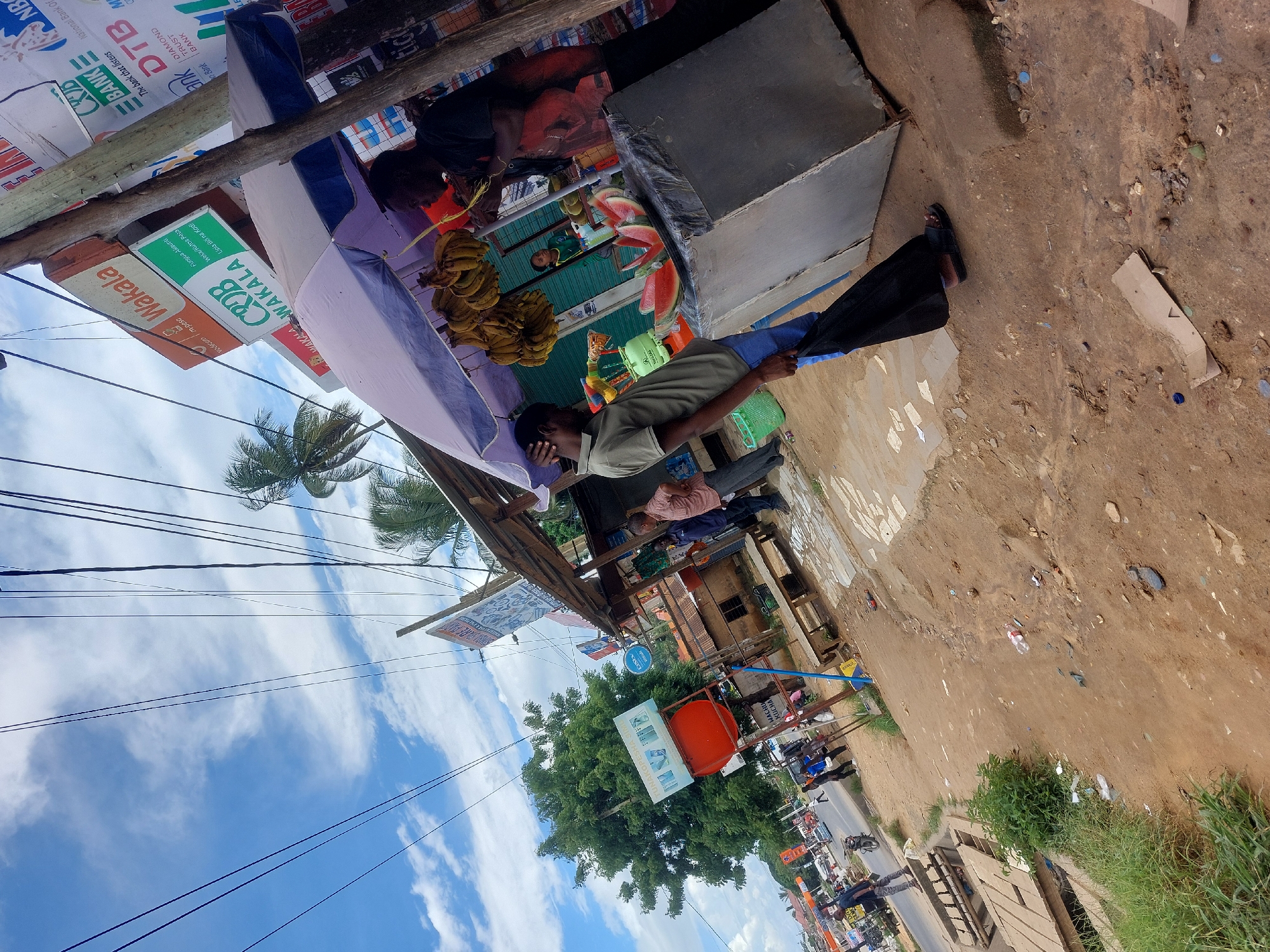 The image shows a street scene in Dar es Salaam, Tanzania. A vendor stands at a fruit stand, selling bananas and watermelons. Signs for local businesses and banks, including Vodacom M-Pesa, CRDB Wakala, DTB Bank, and National Bank of Commerce, are visible on the structure. The environment appears to be a bustling marketplace, with power lines overhead and other buildings and businesses visible in the background. The weather seems sunny and bright. Pedestrians and vehicles can be seen on the street.