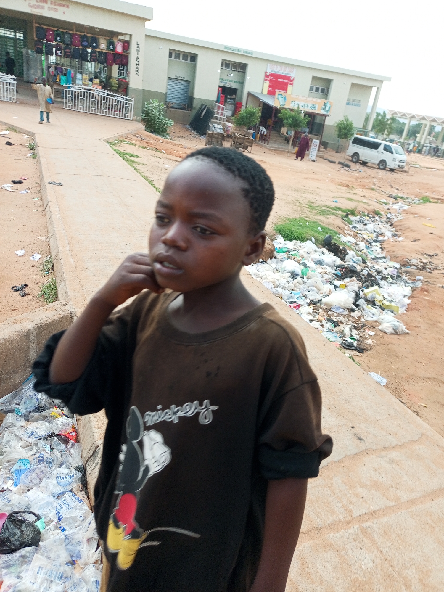 A young boy stands in the foreground of the image, facing to the right. He is wearing a dark brown t-shirt with a graphic of Mickey Mouse and the word "Mickey" printed on it. His left hand is raised to his chin, and his eyes are looking off to the side. The background shows a street scene in what appears to be an outdoor market area. There are several buildings with storefronts lining the street. Piles of garbage, including plastic bags and bottles, are visible on the ground to the right of the boy and in the far background. A white minivan is parked on the street. In the far background, a covered walkway with visible columns suggests a larger market complex. The ground is predominantly dirt and sand, with some patches of green grass. The lighting suggests it is daytime. Based on visual cues such as architecture, vegetation, and the presence of a minivan, the location is likely Gombe, Nigeria.