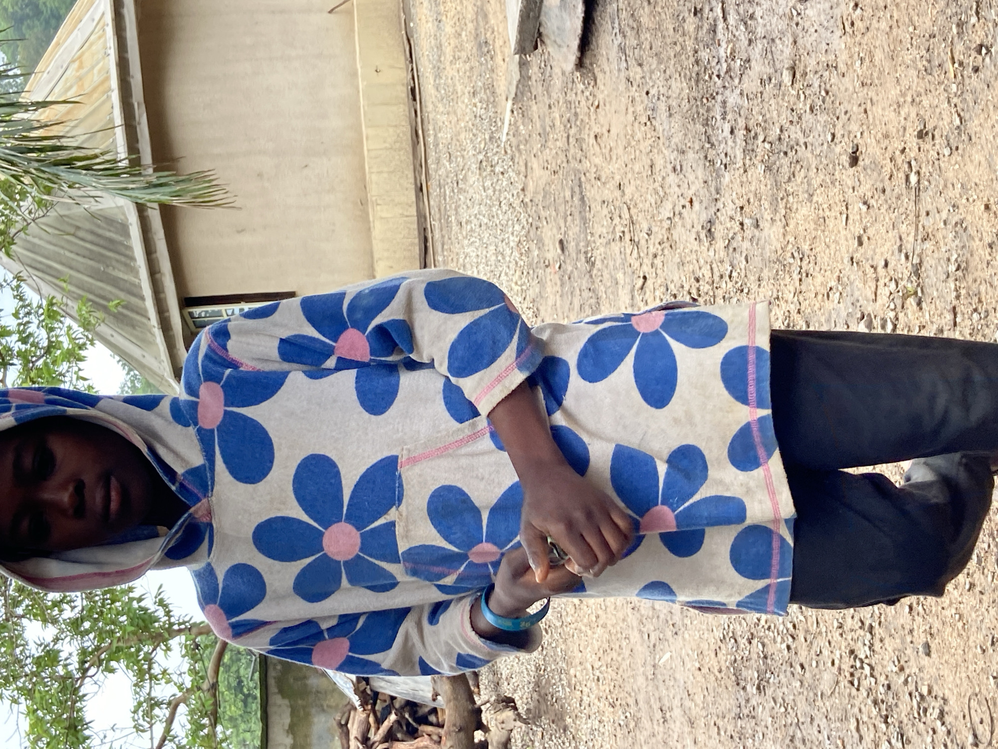 This image, captured in Mararaba, Nigeria, depicts a young individual standing on unpaved ground composed of dirt and gravel. The person, positioned slightly right of the camera's center, wears a hooded sweatshirt patterned with large blue flowers and pink centers over a white background, and dark trousers. The hood is up, revealing a pink interior lining. A light blue bracelet is visible on the left wrist. Both hands are clasped in front, holding a small, unidentifiable light-colored object. In the background, to the left, a pile of firewood rests beneath green tree foliage. To the right, a light-colored building with a corrugated metal roof and a visible window is partially obscured by a hanging palm frond.