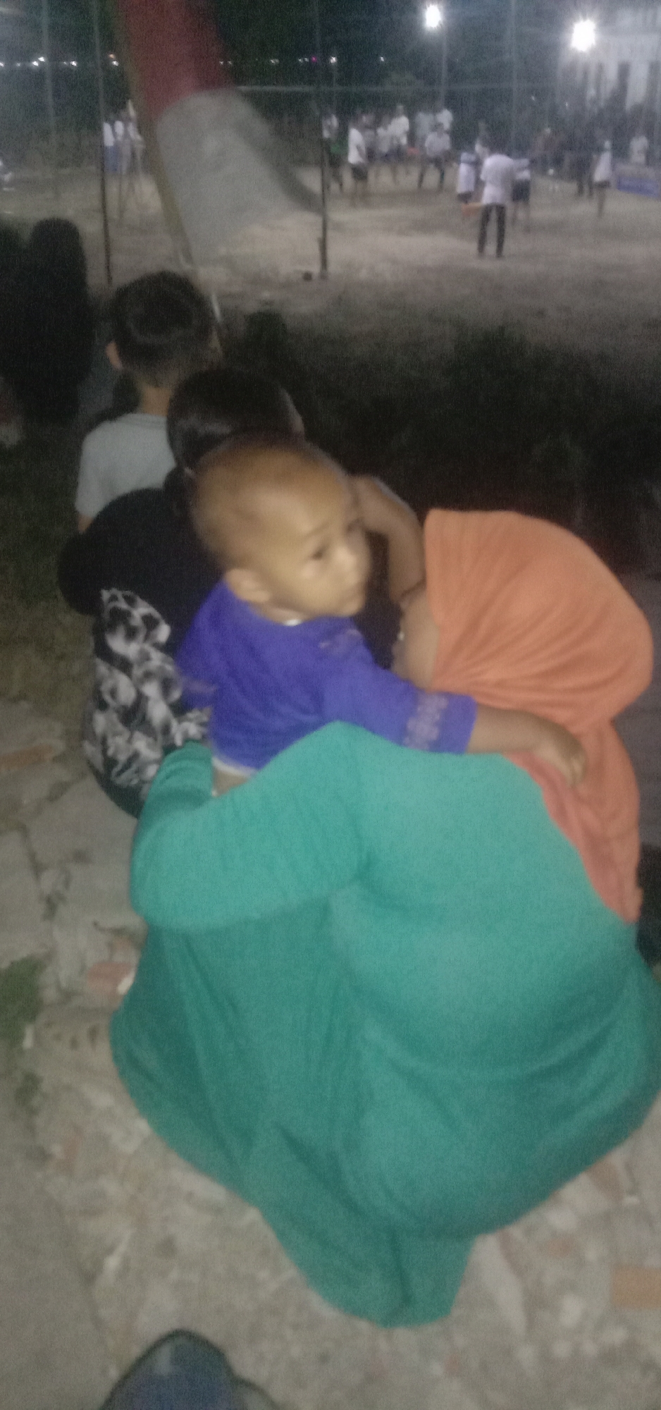 In Banjar Dowo, Indonesia, at an outdoor event possibly in the evening, a young child in a purple shirt is being held by an adult wearing an orange headscarf and a teal garment. The child is looking to the right, with their head tilted. Behind them, another person is visible wearing a black and white patterned shirt. In the background, people are gathered on a sandy or dirt field, with some standing and others appearing to be participating in an activity. Lights are visible in the distance, suggesting the presence of streetlights or floodlights. There is a large, blurry red and white object in the upper left quadrant, possibly a flag or banner. The ground is uneven and appears to be made of dirt and small stones. The overall image is blurry due to low light conditions and the movement of the subjects.