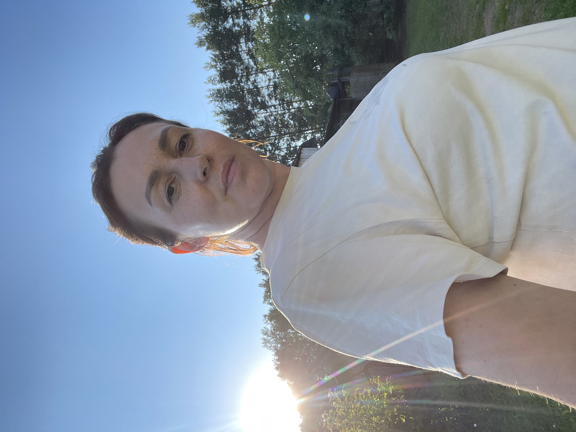 A woman is taking a selfie outdoors in front of a clear blue sky and a line of trees. She is wearing a light-colored t-shirt. The sun is visible on the left side of the frame, casting a flare across the image. The background shows a rural landscape with some wooden structures and green grass. The location is not Bormist, Russia.