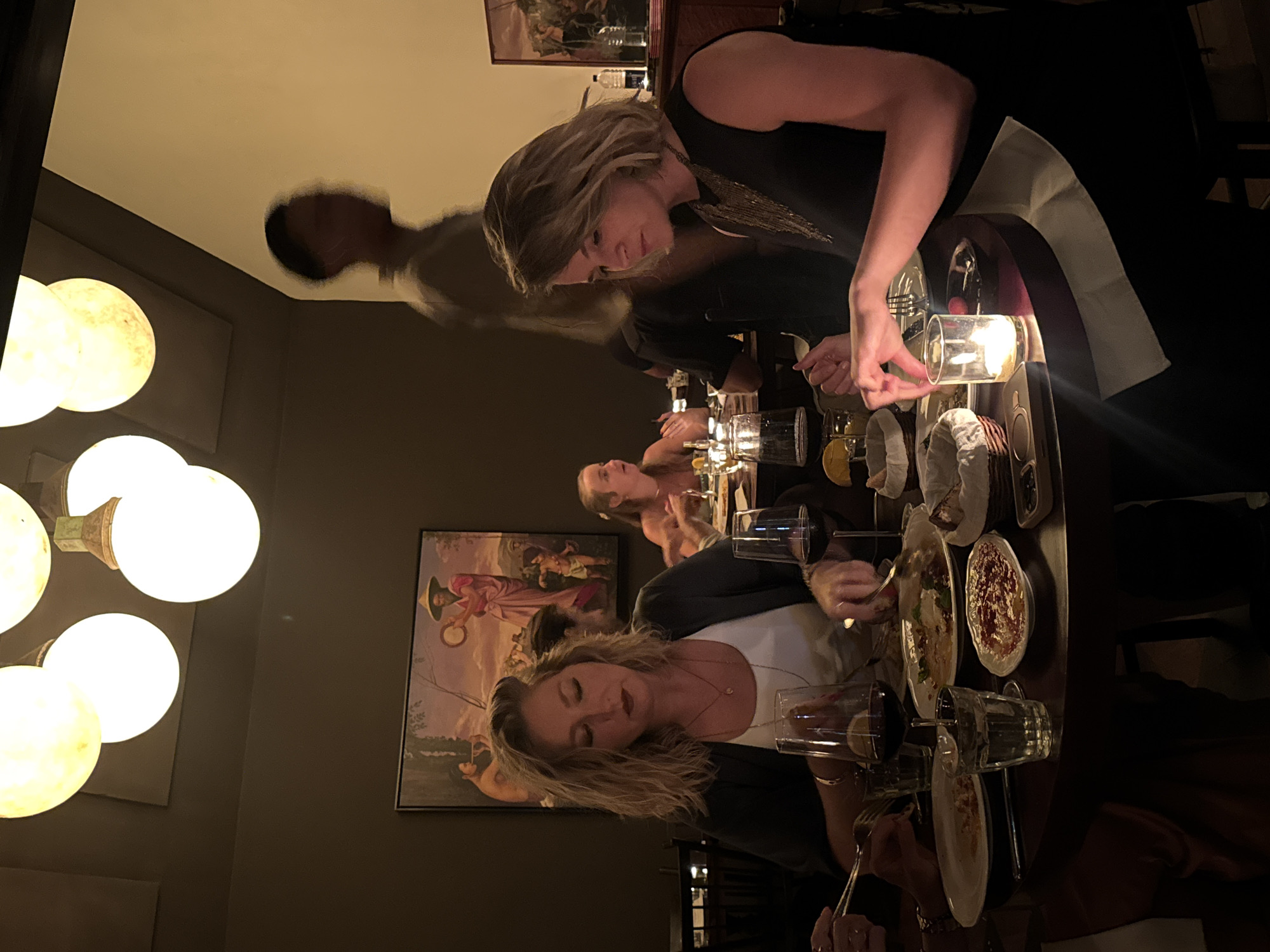 The image captures a dim-lit dining scene, likely in a restaurant or private dining room, featuring several women gathered around a round table. The setting appears to be evening or nighttime, illuminated by warm artificial light from spherical wall-mounted lamps and a candle on the table.
In the foreground, a woman with light, wavy hair, wearing a white top and dark jacket, is focused on her plate, holding cutlery, her head tilted slightly. Reflected in the surface above, a blurred figure of a person is visible. In the mid-ground, another woman with light hair and a dark sleeveless top, adorned with a distinctive golden, layered necklace, leans over the table, seemingly reaching for something near a lit candle in a glass holder. Partially visible further back are other diners at the same table.
The table is set with multiple plates containing food, possibly dips or small dishes, along with wine glasses, water glasses, and cutlery. The ambiance is intimate and social. The walls are dark-colored, featuring at least two framed paintings. The more prominent painting depicts a scene with multiple figures in what appear to be historical or traditional attire. There is no visible text in the image. The overall atmosphere suggests a relaxed, upscale dining experience.