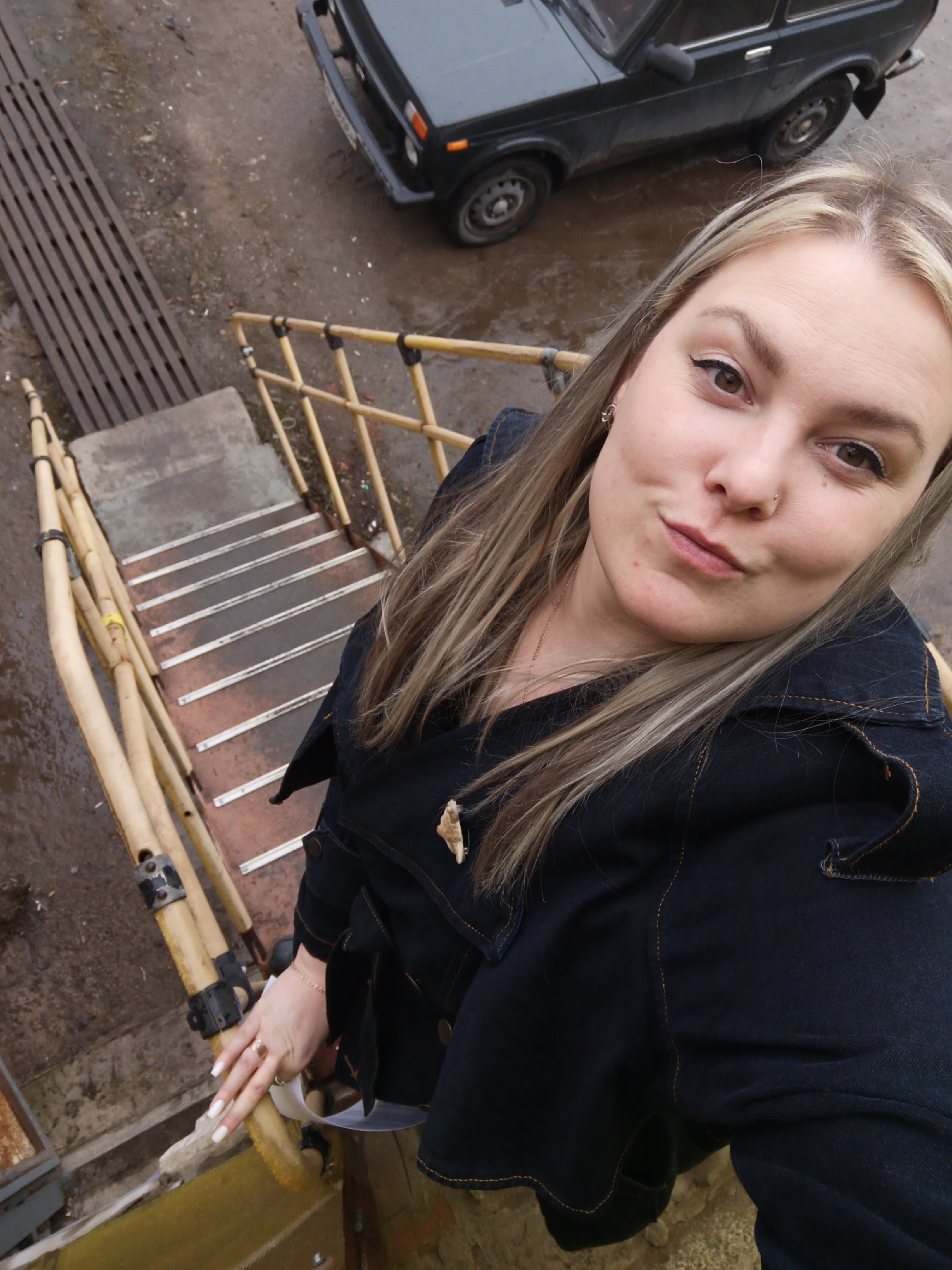 A woman with blonde hair and a denim jacket is taking a selfie from a high angle, standing on a set of stairs with yellow railings. The stairs are painted in alternating shades of gray and orange. The woman is looking at the camera with a slight smirk.
In the background, there's a dark grey or black car parked on a wet, asphalt surface. To the left of the stairs is a metal grate. The image appears to have been taken in an industrial or urban area in Noviy, Russia, as specified.