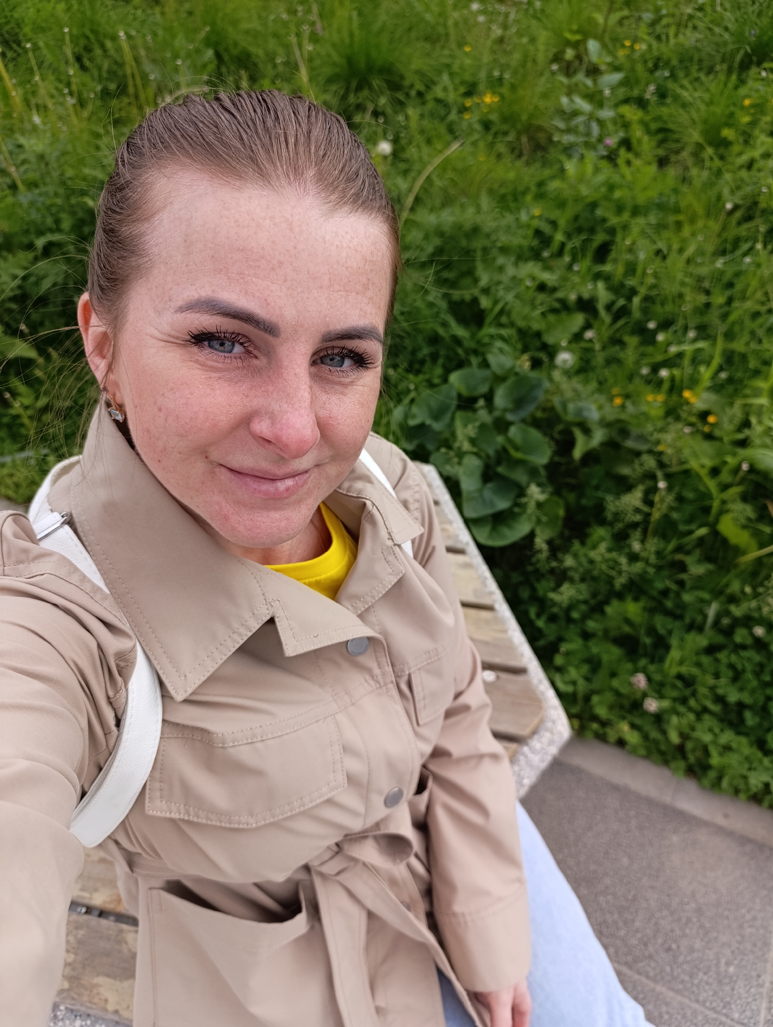 A woman with freckles and blue eyes is sitting on a wooden bench, likely outdoors given the green foliage in the background. She is wearing a beige trench coat over a yellow shirt and has a white strap across her shoulder, possibly from a bag. Her hair is pulled back. The location is Unknown, Unknown. The scene appears to be a selfie or photo taken by the subject.
