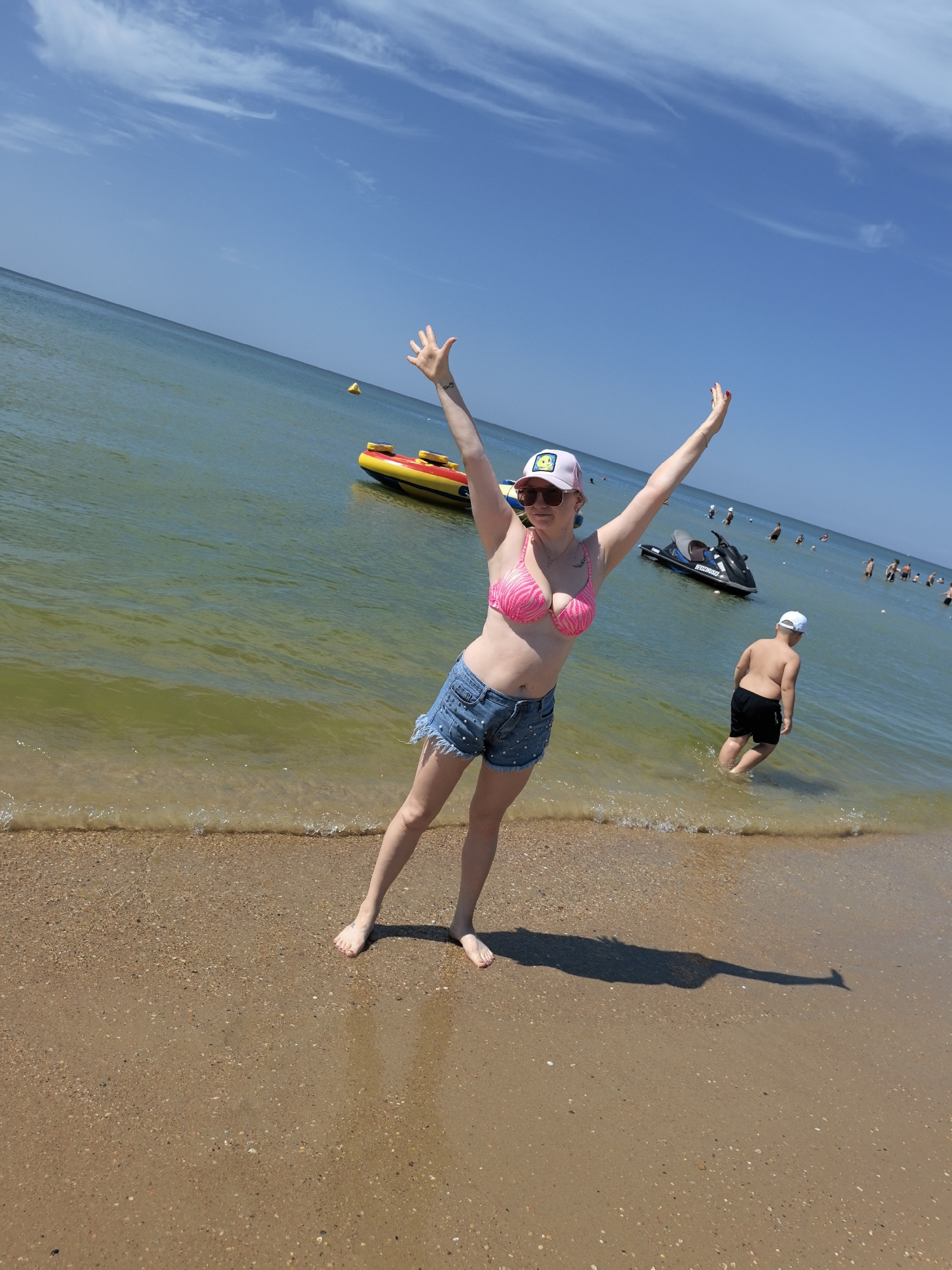 A full-shot, eye-level, outdoor, daytime, vertical, slightly angled, full body, medium-close up photograph shows a young Caucasian woman standing on a sandy beach on the coast of the Black Sea in Anapa, Russia. She is facing forward and slightly to her right, with her arms raised upwards and outwards, her left leg slightly bent and extended forward, and her right leg bent and behind her left. She is wearing a pink and white striped bikini top, denim shorts with frayed edges, a white baseball cap with a yellow smiley face design, and sunglasses. Her expression is happy, with her mouth open and a slight smile. The water is a greenish-blue color, with small waves lapping at the shore. In the background, there is a yellow and red inflatable boat, a black jet ski, and several other people swimming and standing in the water. The sky is clear and blue with a few white clouds. The sun is shining brightly, casting a shadow of the woman on the sand to her right.