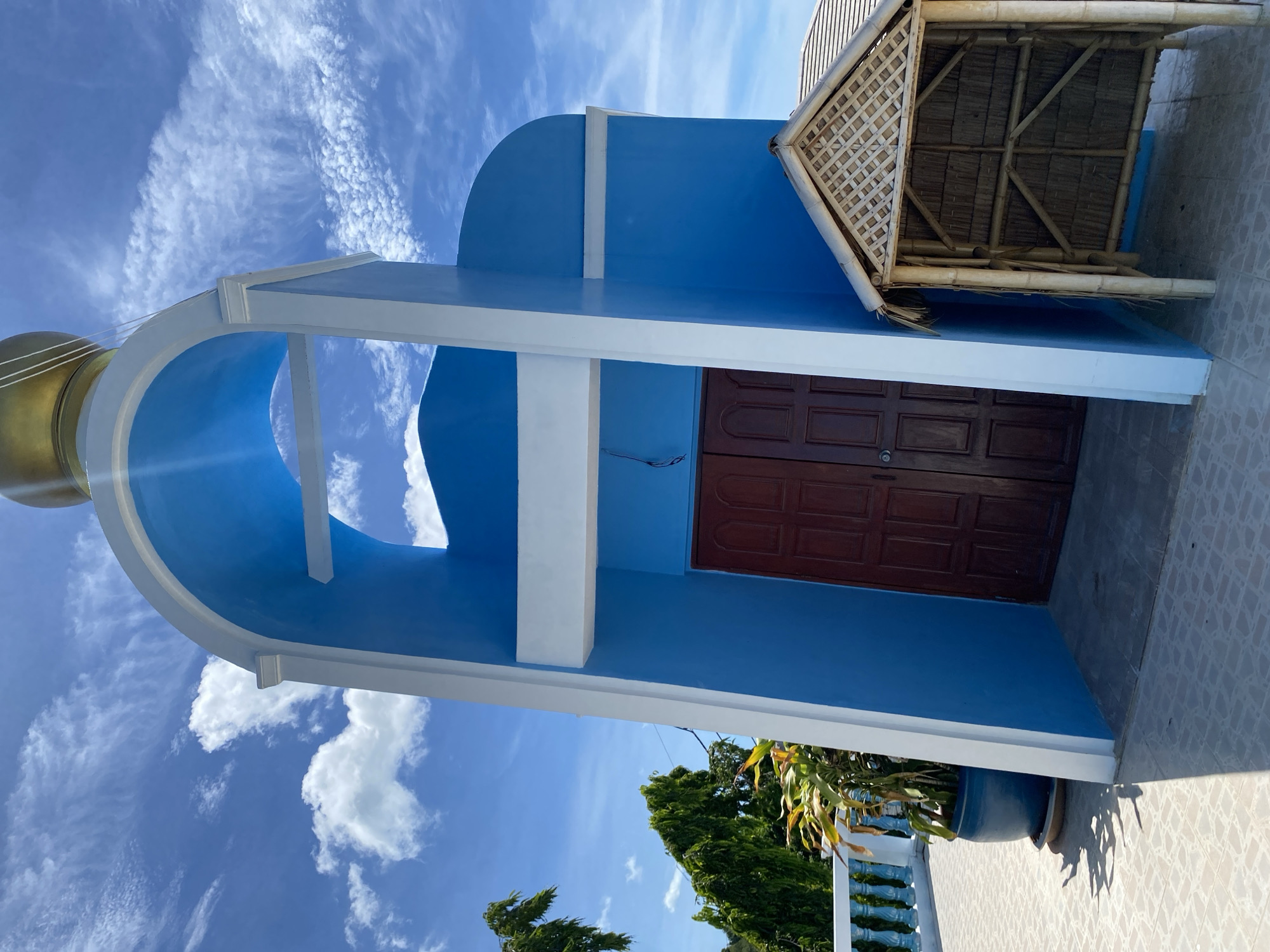A blue and white structure, likely a bell tower or small chapel, stands against a bright blue sky with scattered white clouds. The structure has an arched opening with a golden dome at the top. To the right of the entrance, there is a small thatched-roof structure made of bamboo. In the foreground, there is a tiled patio. To the left of the main structure, a potted plant sits on the patio, and beyond it, a white railing and lush green trees are visible. The scene appears to be in a tropical environment. The location is identified as Baan Hua Thanon, Thailand. There are no people visible in the image.