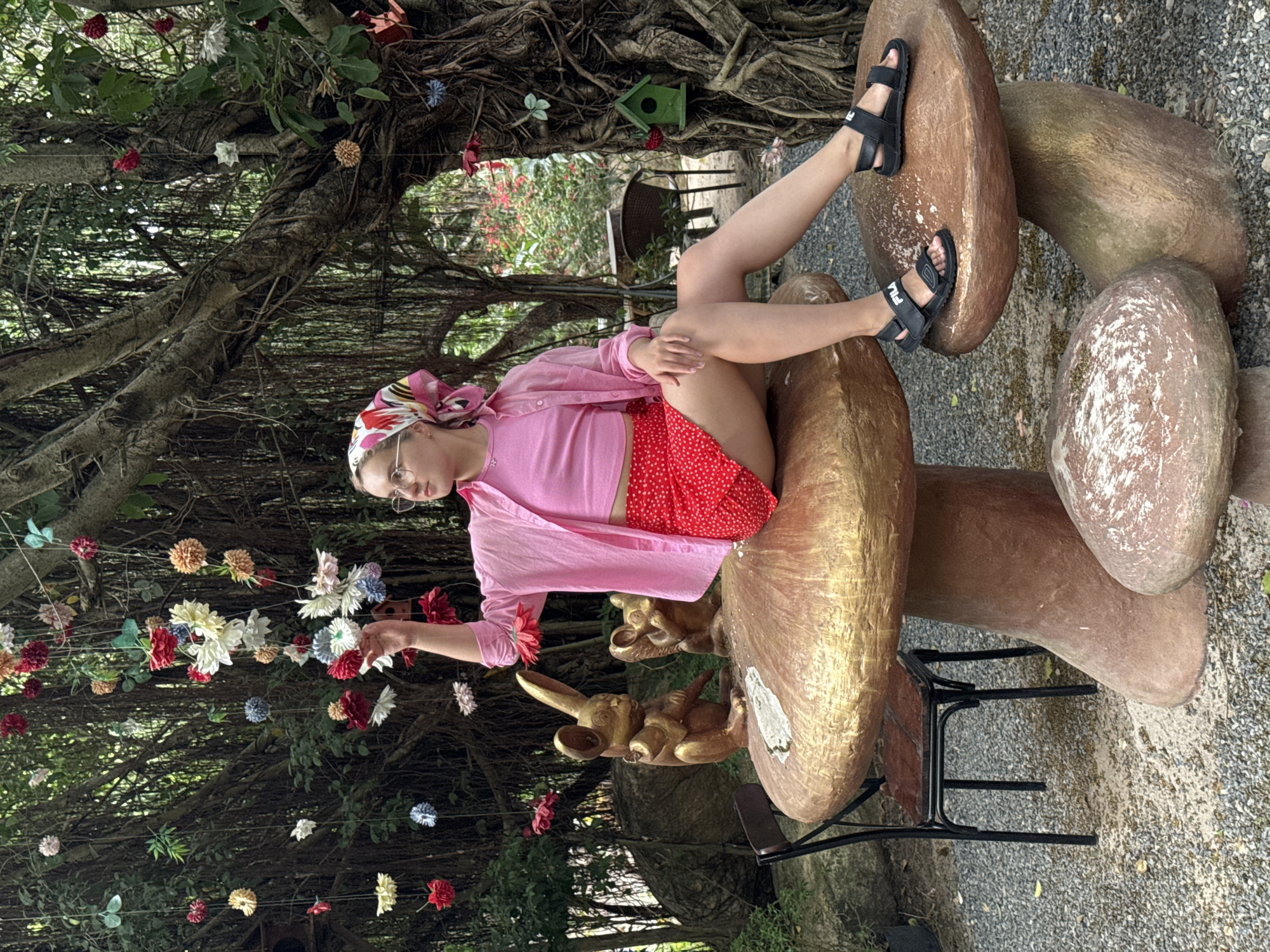 A woman is reclining on a cluster of oversized mushroom sculptures, in what appears to be a whimsical garden setting, possibly in or near Nam Nha Trang, Vietnam. The woman is wearing a pink headscarf, round glasses, a pink crop top, a pink button-down shirt, and a red polka-dot skirt. She is wearing black Fila sandals. The mushroom sculptures are painted in shades of brown and gold and are positioned on a black metal chair frame. Underneath the mushroom is a rabbit sculpture in gold. Above the scene is a banyan tree with aerial roots and hanging decorations of multicolored paper flowers. The ground is covered in gravel. In the background is a lush green area with a birdhouse.