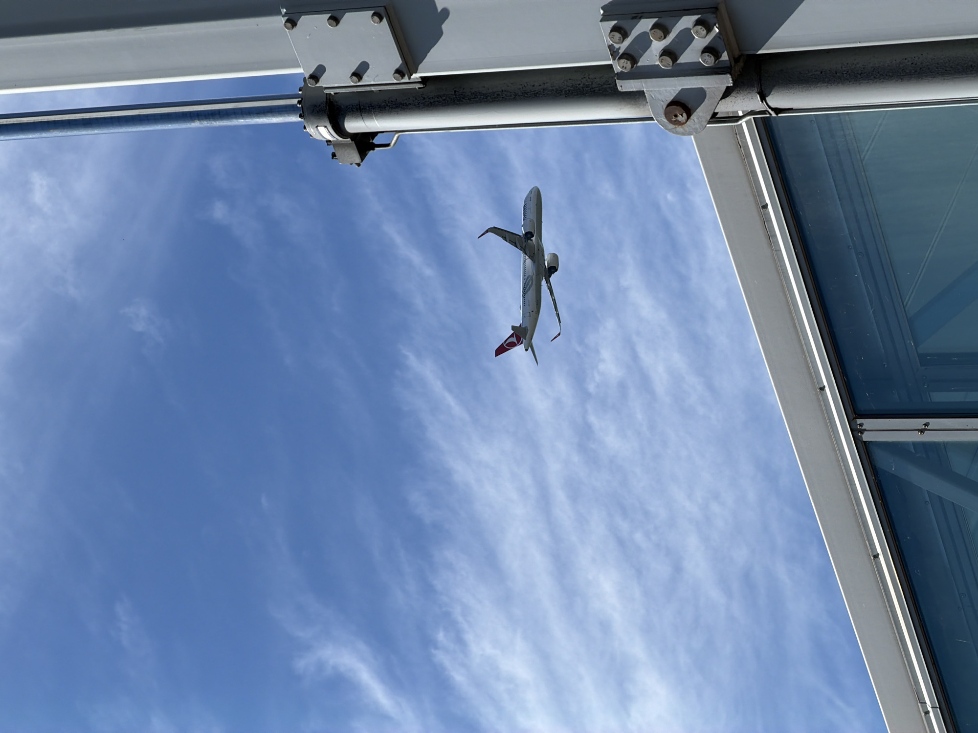 An airplane with the Turkish Airlines logo on its tail flies through a blue sky with wispy clouds. The airplane is in the upper left quadrant of the image. In the foreground on the right, a large metal structure, part of an airport jet bridge, with bolts and seams is visible, obscuring the lower right quadrant of the image. The lower left quadrant shows the glass exterior of the jet bridge. The image was taken in Tallinn, Estonia.