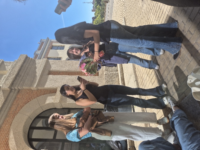 The image depicts five individuals and a partial sixth person standing or reclined outdoors on a sunny day. The scene is situated on a light brown paver-covered ground in front of a multi-story building with a light-colored stone facade and reddish-brown brick arched window frames. The sky is clear blue, and strong sunlight casts distinct shadows on the pavement.
Four young women are standing:
1. On the left, a woman with light brown hair wears a light blue short-sleeved top and white wide-leg trousers, carrying a brown jacket over her left arm.
2. In the center-left, a woman with dark hair wears a black tank top and dark blue flared jeans, actively looking at a smartphone held in her hands.
3. In the center-right, a woman with dark hair wears a black top and dark blue flared jeans, holding a large bouquet of pink and green flowers and smiling towards the right.
4. On the right, a woman with long dark hair wears a black short-sleeved top and dark blue flared jeans, positioned with her back mostly to the camera, facing the woman holding the flowers.
In the lower-left foreground, the legs of a fifth person are visible, clad in blue jeans and white sneakers, extended as if seated or reclined on the ground. A light-colored bag is positioned near these legs. On the far right, a hand holding a smartphone extends into the frame from a partially visible sixth individual. A metal mesh trash receptacle is visible in the mid-ground to the left. The architectural style and outdoor setting are consistent with the campus of the Universidad Complutense de Madrid, Spain.