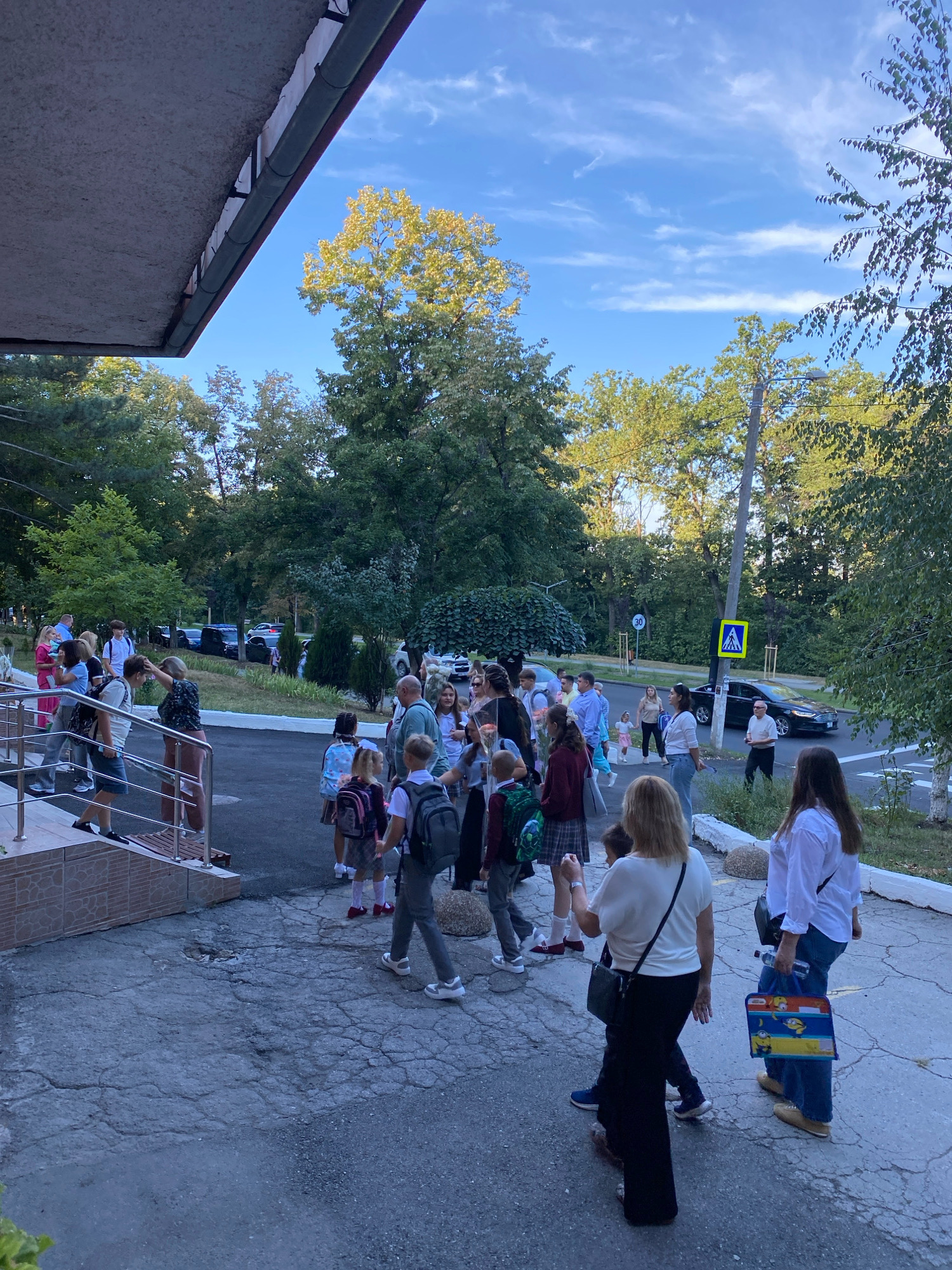 The image depicts a group of people, likely students and adults, gathered outside a building that appears to be a school, possibly on the first day of the academic year, given the presence of backpacks and flowers. The setting is outdoors with mature trees and a clear blue sky with wispy clouds, suggesting a pleasant morning or afternoon in Chișinău, Moldova. The architecture visible in the foreground suggests a building with an overhang and stairs leading up to an entrance. People are seen walking, standing, and interacting, with some holding backpacks and others carrying bags or bouquets of flowers. The overall atmosphere appears to be one of community and anticipation.