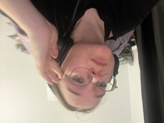 A young female, likely Caucasian, is depicted indoors in Pushchino, Russia. She wears round, thin-framed eyeglasses and dark over-ear headphones. Her hair, appearing dark, is partially visible. She is dressed in a dark, short-sleeved top and a thin silver-colored chain necklace. Her left hand, adorned with a thin ring on the ring finger, is raised to the left side of her face, with fingers near her temple and ear. She exhibits a neutral facial expression while looking directly at the camera. The background consists of a light-colored wall, a dark curtain on the far left, and blurred objects in the mid-ground, including a purple fabric item and other indistinct articles.