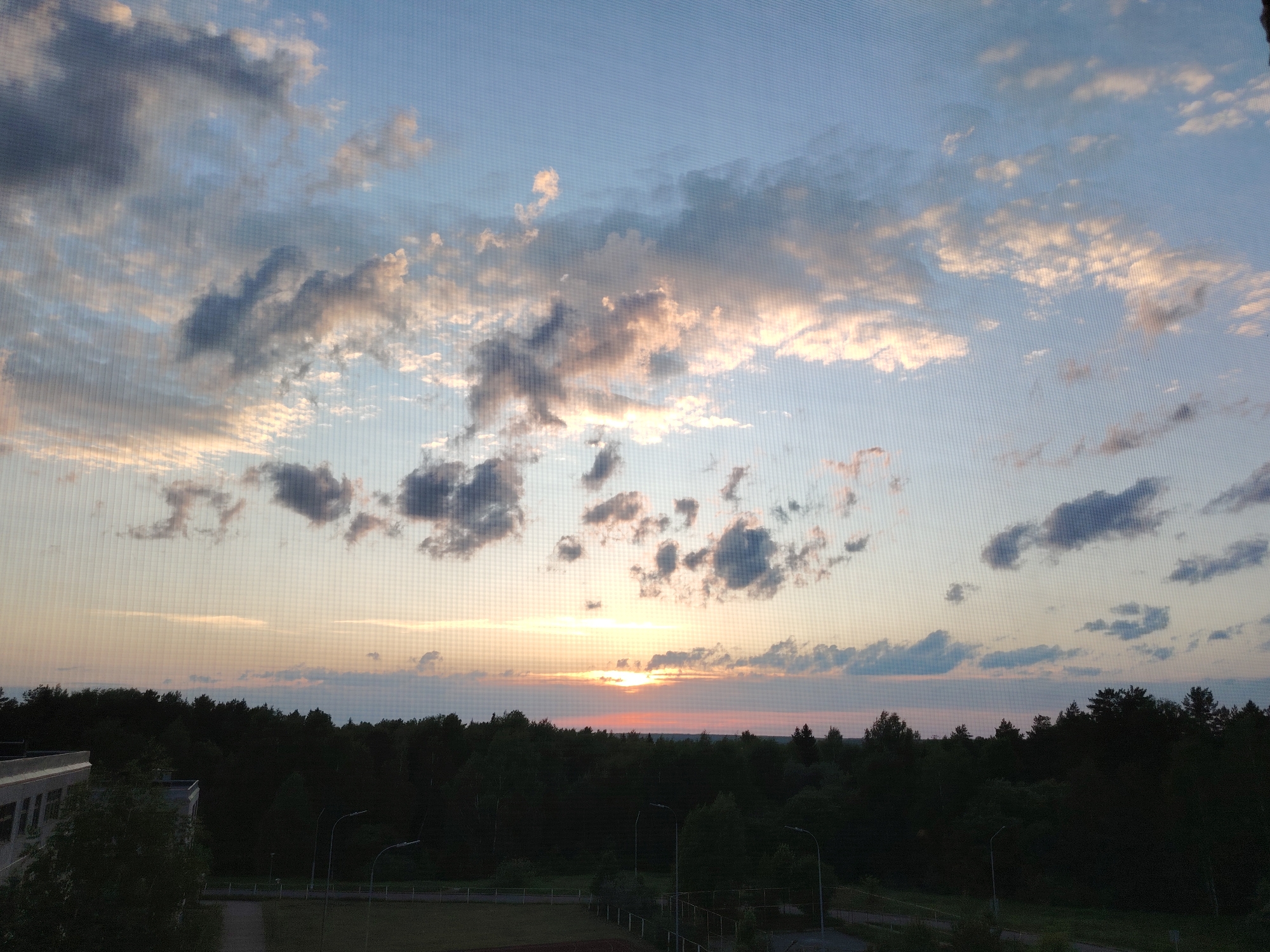 The image shows a sunset scene in what appears to be Марковский, Russia. The sun is setting on the horizon behind a dense forest, casting a warm glow across the sky. There are scattered clouds in the sky, some illuminated by the setting sun, others appearing darker. In the foreground, there's a building on the left and a sports field with running tracks. Streetlights are visible among the trees. The photo seems to be taken through a window screen.