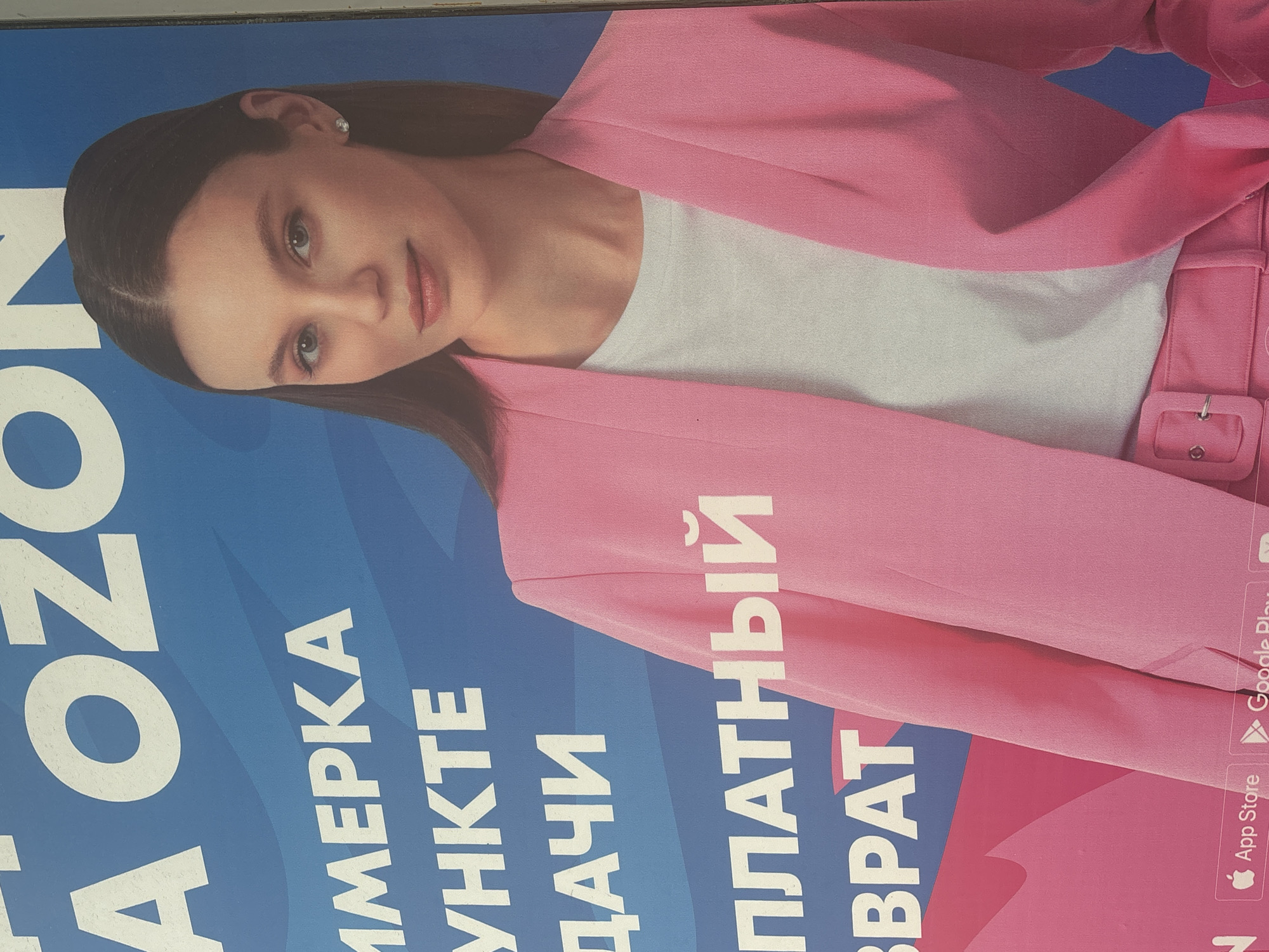 The image is an advertisement featuring a young woman in a pink blazer. The advertisement is likely for an online retailer, possibly Ozon, as suggested by the text "OZON" visible at the top of the image. The woman is looking directly at the camera with a neutral expression. The background is a blue gradient with white text that appears to be in Russian.
The question about the location in the city of Novosibirsk, Russia, cannot be answered from the provided image. There are no visual cues or text within the image that indicate a specific city or location. The image itself is a close-up of a poster or advertisement, and does not depict an outdoor scene or any recognizable landmarks.