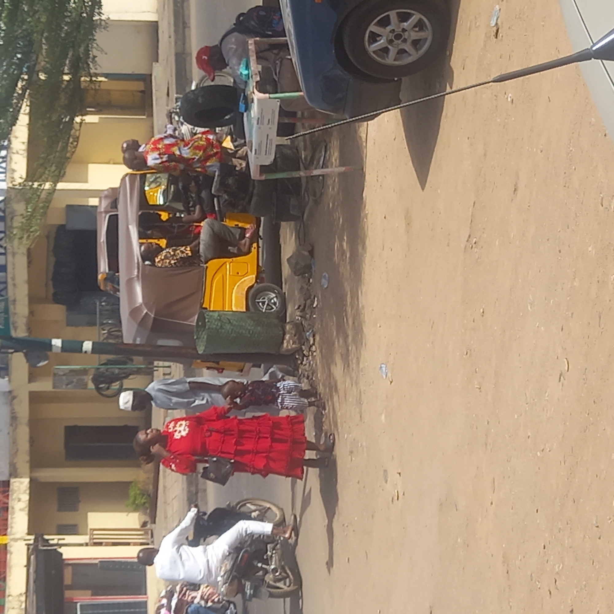 This daytime photograph captures a bustling street scene, likely in an urban or semi-urban area of Dadi Riba, Nigeria. Strong sunlight indicates a clear day, with distinct shadows cast on the light-brown, sandy ground.
In the foreground, a woman dressed in a striking red, ruffled gown, holding a handbag, stands on a dirt path or sidewalk. A child in a striped shirt stands close to her. Nearby, a man wearing a white kufi (cap) and a light-colored shirt is visible. To the right, another man in a white tunic (agbada) is seated on a motorcycle.
The middle ground features a yellow auto-rickshaw, often called a "keke napep," which appears to be parked or moving slowly. A person wearing a patterned shirt is visible inside. A dark blue car is partially seen on the right, with a man in a red cap standing near it. Adjacent to the car is a makeshift stall or table, possibly displaying goods for sale, and a large tire rests on the ground. Green, cylindrical objects, possibly bins or planters, are also on the sidewalk.
Buildings with light-colored facades line the street in the background, suggesting commercial or residential structures. Green foliage from trees is visible on the far left, adding to the urban landscape. The image depicts typical daily life, with people engaged in movement and commerce along a busy thoroughfare. No legible text is visible in the frame.