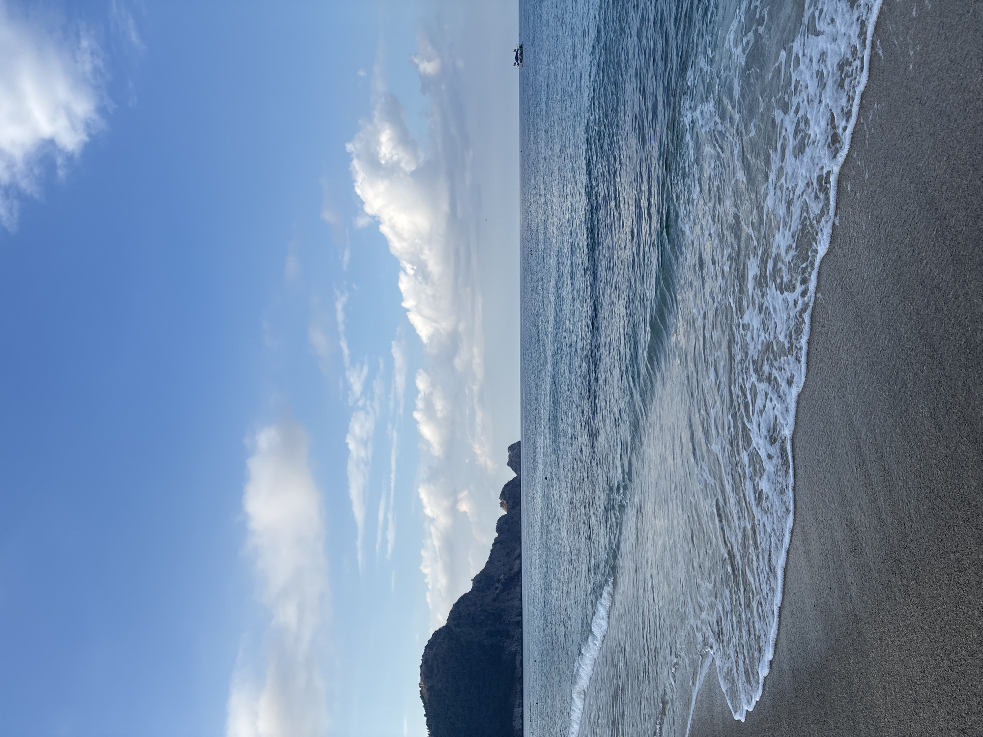 The image depicts a serene seascape with a clear blue sky dotted with white clouds, stretching over a calm blue ocean. The foreground shows a sandy beach with gentle waves lapping at the shore. In the distance, a dark, tree-covered mountain range with rocky formations juts out into the sea on the left side of the frame. A small boat with flags is visible on the horizon on the right. The overall scene suggests a peaceful coastal environment, likely in Alanya, Turkey, known for its beautiful beaches and mountainous coastline.