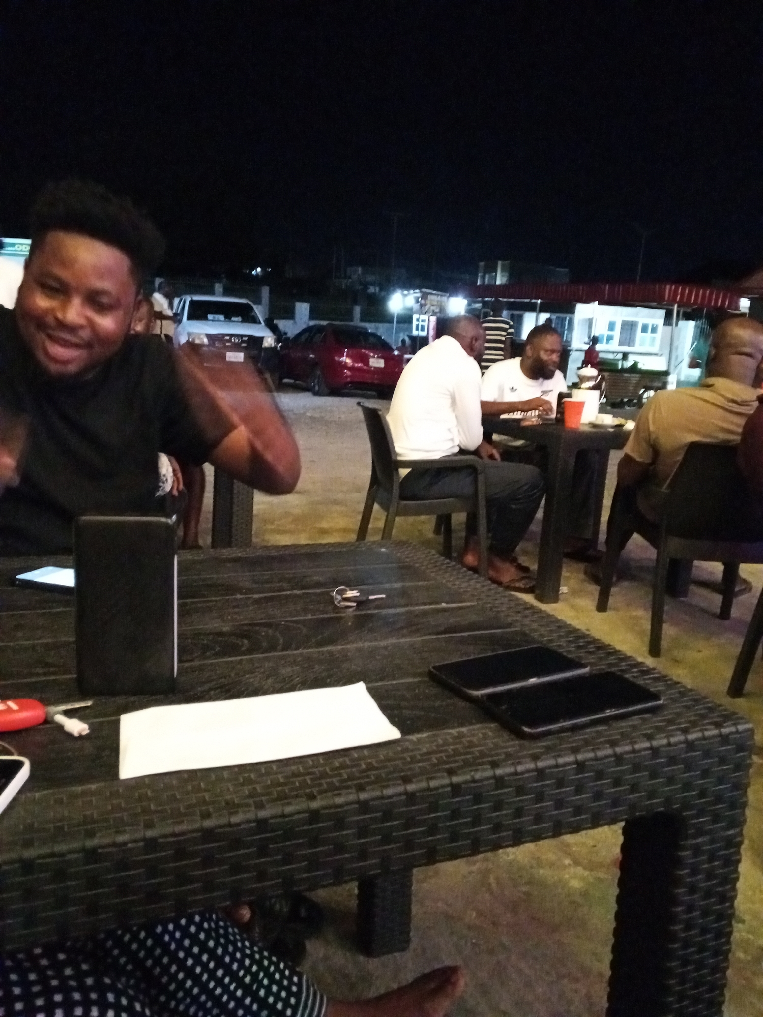 Four African men are seated at two black wicker tables at an outdoor venue at night. At the first table, one man is seated with his back to the viewer, and another man is wearing a white t-shirt. At the second table, one man is smiling and gesturing with his hands, and the viewer can see part of another person’s patterned clothing under the table. On the second table is a rectangular block, a piece of white paper, a set of keys, and two black mobile phones. A white pickup truck and a red car are parked nearby, and some buildings are visible in the background under the night sky. The location is Lokoja, Nigeria.