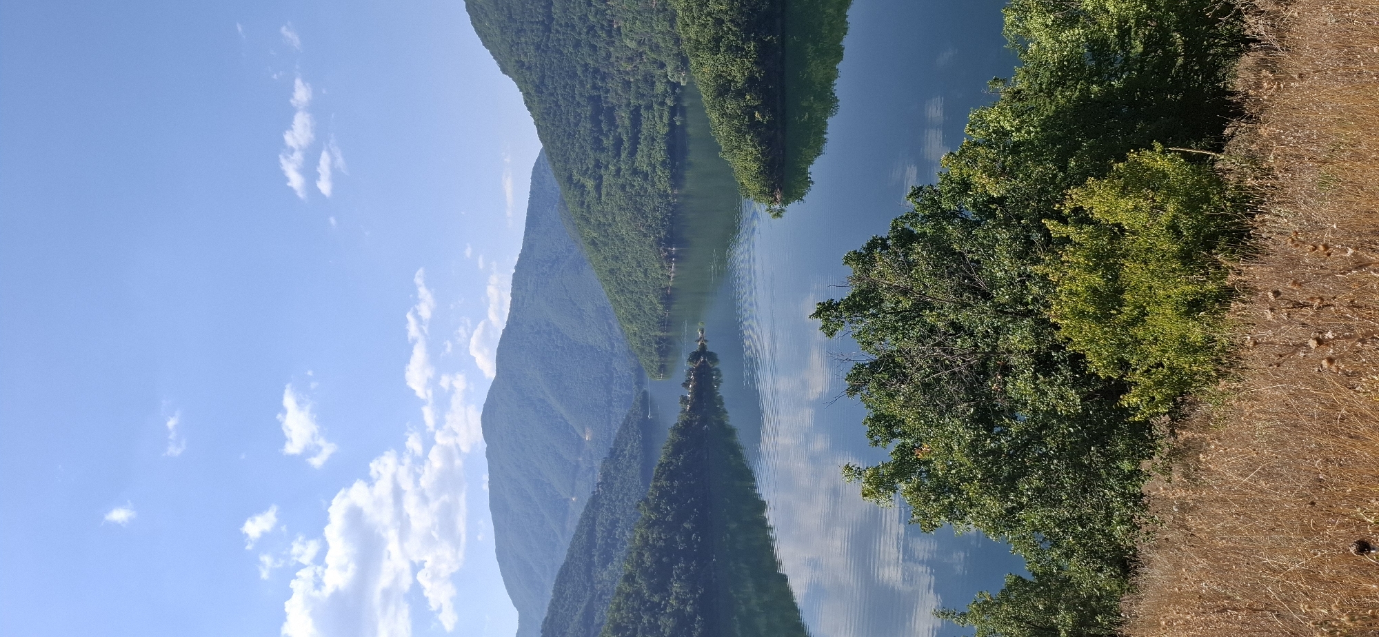 The image depicts a scenic landscape featuring a wide, calm body of water surrounded by lush green hills and mountains. The sky is a vibrant blue with scattered white clouds. In the foreground, dry, brown vegetation covers the ground, transitioning into dense green foliage further up the slope. The water reflects the sky and the surrounding greenery, creating a serene and picturesque scene. The location is identified as Mihalkovo, Bulgaria. There are no people or discernible human activities visible in the image.