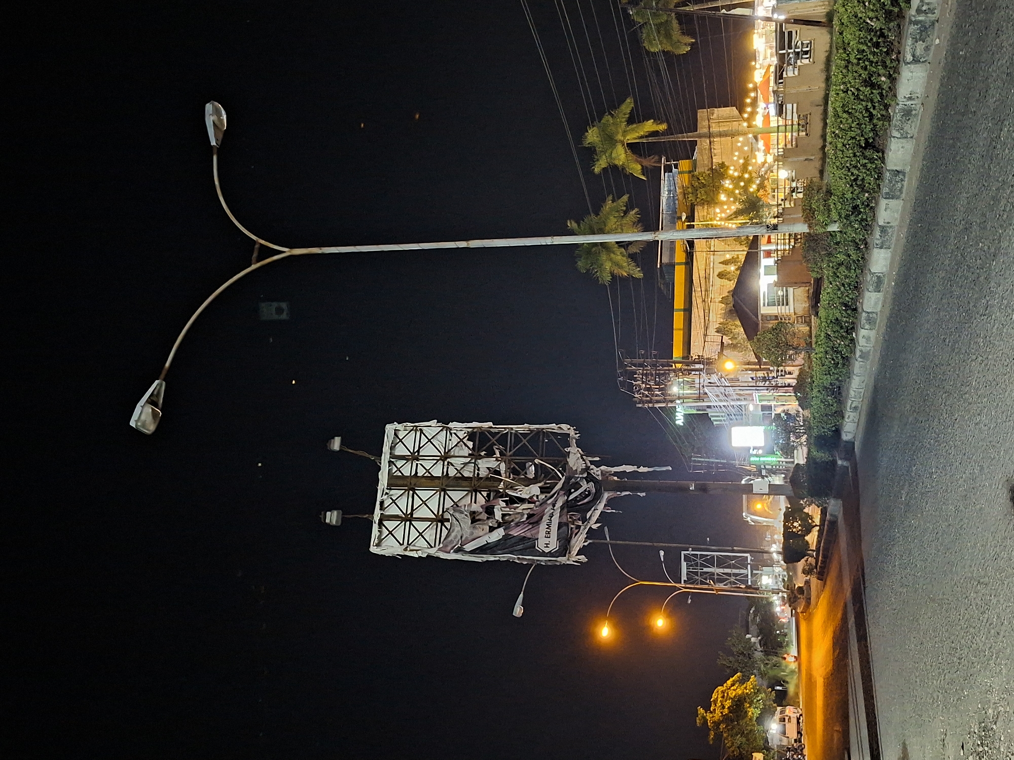 At night, a street scene in Lhokseumawe, Indonesia, reveals a damaged billboard with tattered remains of an advertisement featuring the name "H. ERMIAN". The billboard's structural framework is visible, illuminated by surrounding streetlights. Overhead power lines stretch across the sky, and nearby palm trees add to the tropical urban landscape. Along the right side of the image, a section of road with a curb is visible, lined with hedges. Further down the street, bright lights from commercial establishments and passing vehicles illuminate the scene. The overall ambiance is a mix of urban lighting and the darkness of night.