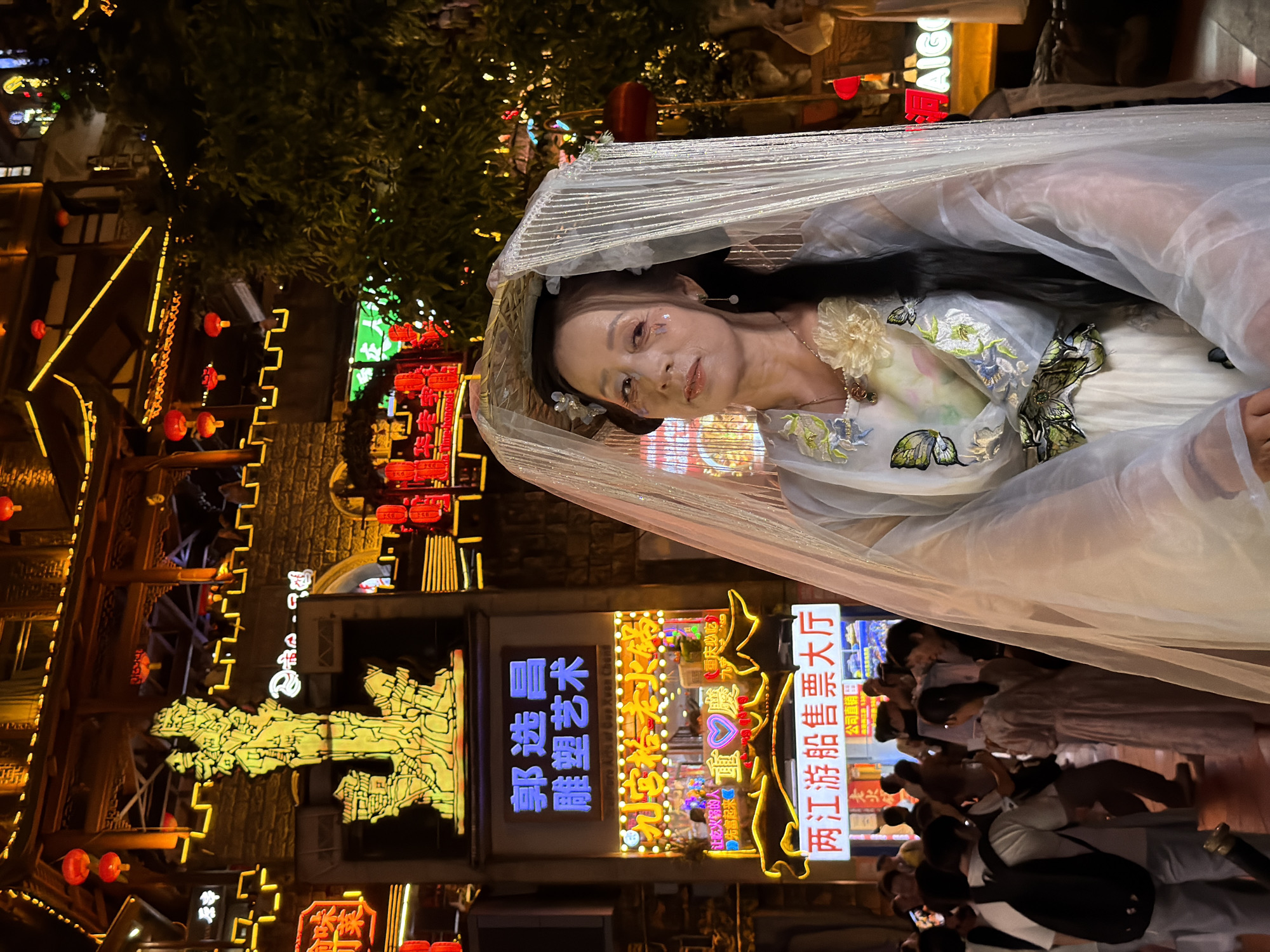 A woman in traditional Chinese attire, adorned with a veil and intricate floral embroidery, is the central subject of this nighttime photograph. She is positioned within a bustling street scene illuminated by an abundance of neon signs and red lanterns, typical of a commercial area in Yuzhong District, China. The woman's gaze is directed upwards, giving an impression of serene contemplation or perhaps posing for the camera. Her makeup features subtle glitter accents on her cheeks and eyes, adding to the ethereal quality of her appearance.
Behind her, a vibrant and densely packed urban environment unfolds. The buildings are adorned with bright, colorful signs, some displaying Chinese characters, indicating shops and businesses. Notably, there are signs for "两江游船售票大厅" (Liangjiang Cruise Ticket Hall), "雕塑艺术" (Sculpture Art), and "重庆必吃" (Chongqing Must-Eat), all rendered in glowing neon. The architecture appears traditional, with dark wooden elements and tiered structures, contrasting with the modern vibrancy of the signage.
A crowd of people can be seen in the background, suggesting a lively atmosphere, possibly a tourist area or a night market. The time of day is clearly night, as indicated by the artificial lighting and the darkness of the sky. The overall mood is festive and engaging, capturing the essence of a lively urban night in China. The composition, with the woman framed by the architectural elements and the vibrant lights, creates a striking visual narrative.