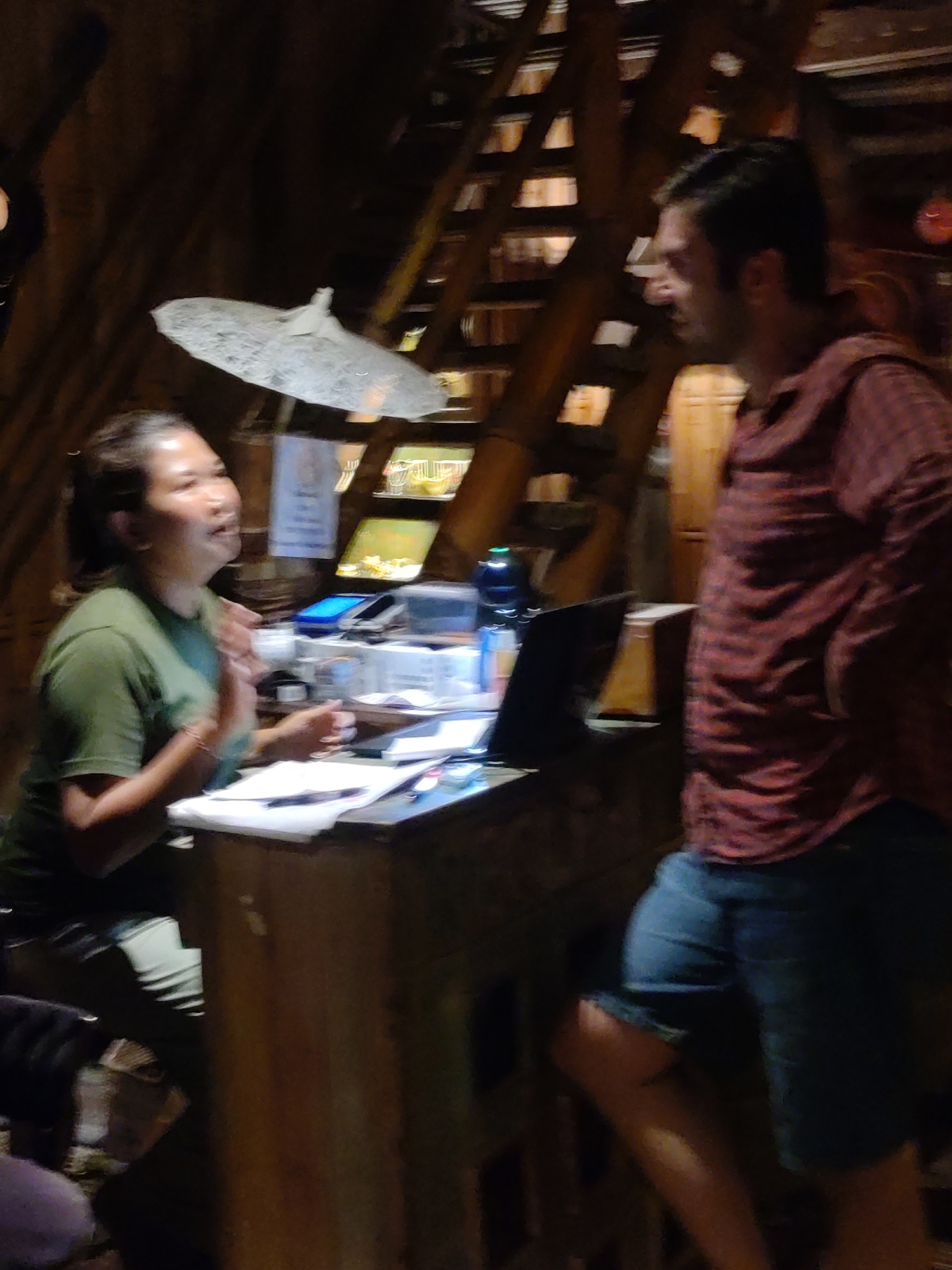 The photo, taken indoors in Lokaserena, Indonesia, shows two people engaged in a conversation. A woman in a green t-shirt is seated behind a wooden counter. She is facing left, with her hands in front of her as if gesturing while speaking. The counter contains various items including papers, an open laptop, electronic devices, and bottles. To the right of the counter stands a man wearing a red plaid shirt and blue denim shorts. He is facing the woman and appears to be listening. The background features a wooden structure, possibly part of the building's interior, with a decorative light fixture overhead.