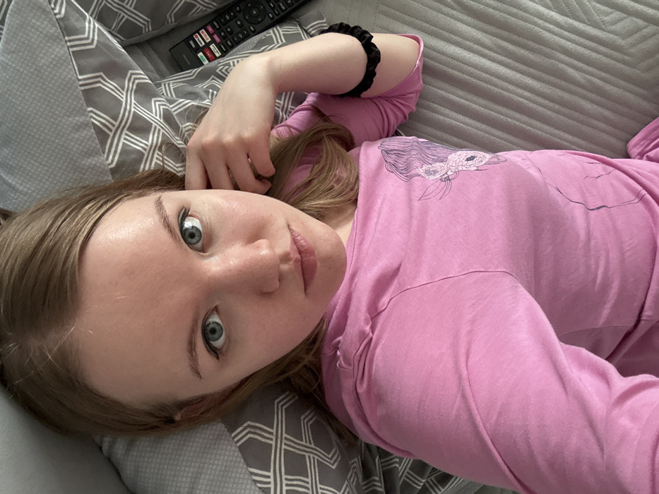 An individual, identified as an adult female, is depicted in a close-up, high-angle shot, lying on her back. She possesses light brown hair, centrally parted, which frames her face and extends onto the bedding. Her eyes are blue with visible black eyeliner on the upper lash line. She exhibits a fair complexion and a neutral facial expression, with her gaze directed towards the camera.
She is wearing a long-sleeved pink top. The top features a graphic design on the left chest area, illustrating a stylized profile of a head with flowing hair and floral elements. A black scrunchie is present on her right wrist. Her left hand is partially visible, resting near her left temple or ear.
The subject is positioned on bedding. Behind her head is a pillow with a grey and white geometric pattern. To her right, a portion of a grey duvet cover or sheet with horizontal stitched lines is visible. A black television remote control, displaying multiple colored buttons (red, green, yellow, blue) and white text including "DVD," is located on the bedding near her head. The environment appears to be an indoor setting.
The specific location in the city of Nizhnekamsk, Russia, cannot be determined from the visual information provided in the image.