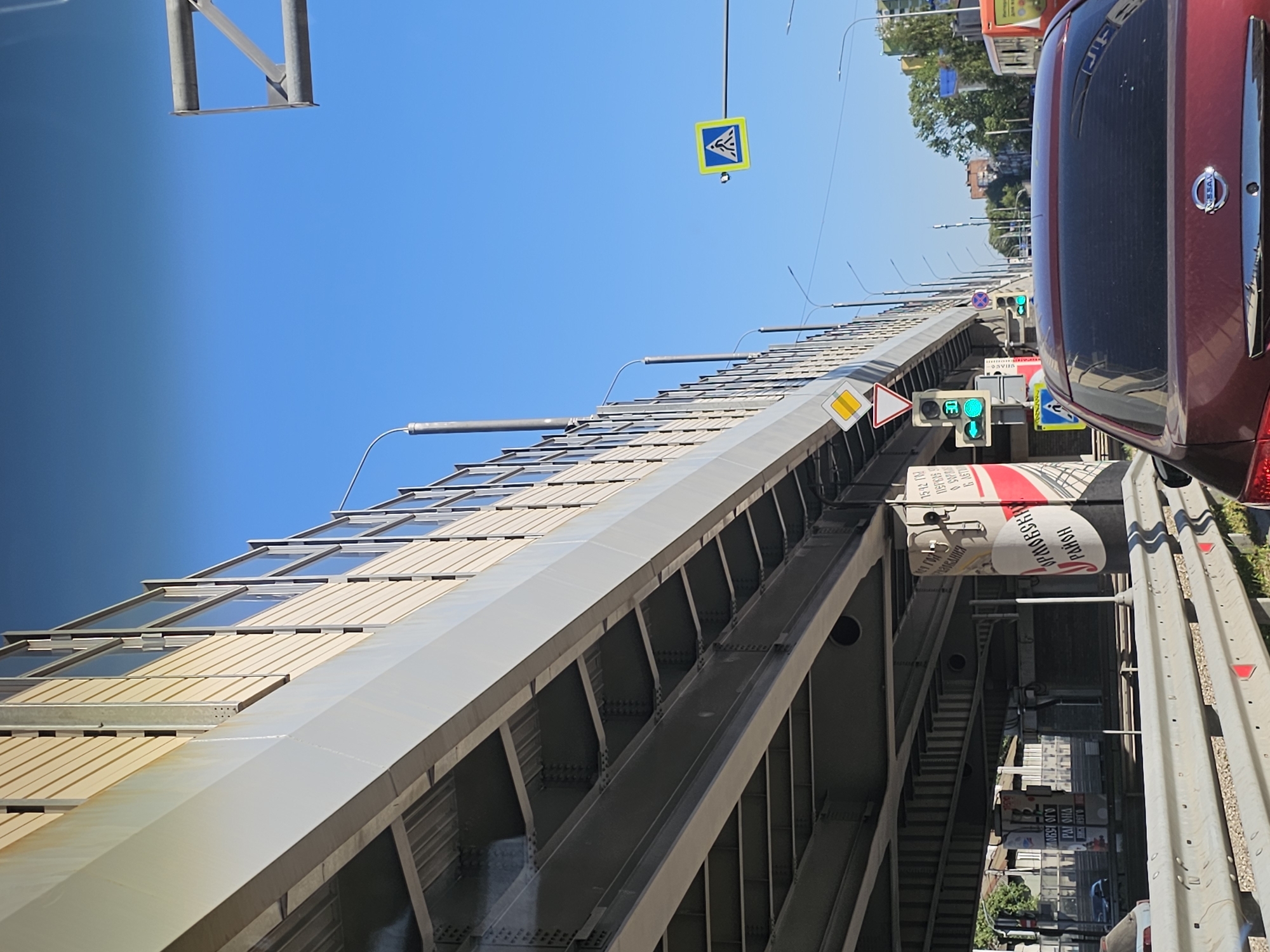 The image depicts a view from a car stuck in traffic under a highway overpass in Nizhny Novgorod, Russia. The overpass, with its beige vertical panels and metal support structure, dominates the left side of the frame, casting a shadow over the road below. The sky is a clear, bright blue.
Traffic lights are visible, showing green signals. A pedestrian crossing sign is mounted on a pole to the right. The cars in the traffic jam include a dark red Nissan sedan in the foreground, its rear visible. On the pillars supporting the overpass, there are large, colorful advertisements. One on the right pillar features red and white text that appears to be in Cyrillic script, including "ОПМОЕВСКАЯ РАЙОН" (Ormoevskaya District). Another pillar further back also has advertisements with Cyrillic text. A metal guardrail separates the lanes of traffic. In the distance, buildings and trees can be seen, suggesting an urban environment.