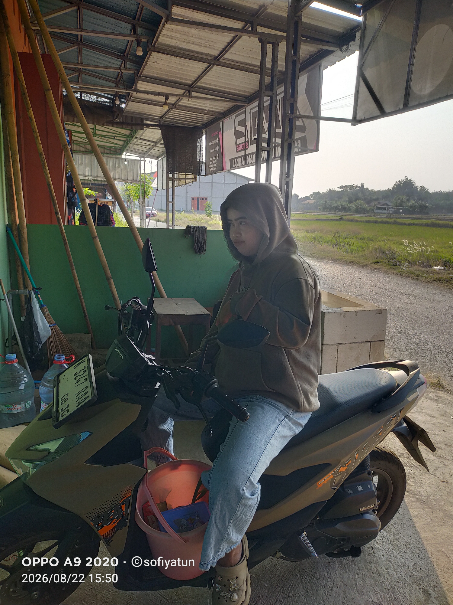 A young woman wearing a grey hooded sweatshirt and blue jeans sits on a dark grey scooter. She is looking to her left, with her arms crossed. The scooter is parked on a paved surface next to a green wall. To the left of the scooter are two large water bottles, a broom, and some bamboo poles. In the background, a rural landscape with rice fields and trees can be seen. The image is timestamped "OPPO A9 2020 - ©sofiyatun 2026/08/22 15:50" and is identified as being from Tayu, Indonesia.