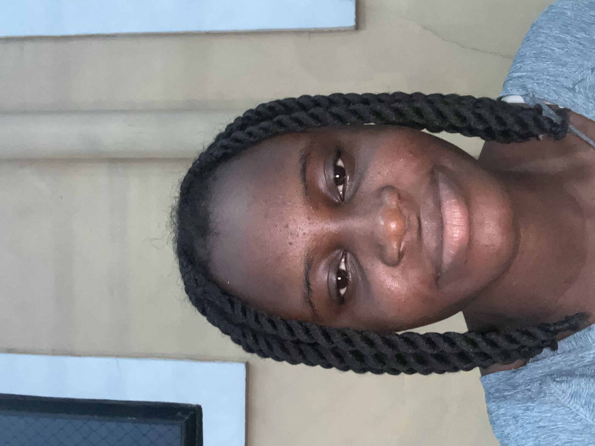 A close-up, head-and-shoulders portrait of a young female is centered in the frame, taken in Ihiagwa, Nigeria. She has dark skin and her dark hair is styled in prominent twist braids that frame her face and extend over her shoulders. She is wearing a light blue-grey, textured long-sleeved top, visible from the upper chest. Her eyes are open, with dark irises, and she gazes directly at the camera. Her expression is neutral with a subtle, closed-mouth smile. The background consists of a beige-tan wall. To the left of the subject, a dark-framed window or opening is visible against a lighter blue background. To the right, a vertical light blue segment appears against the beige wall. The lighting is soft and even, casting no harsh shadows.