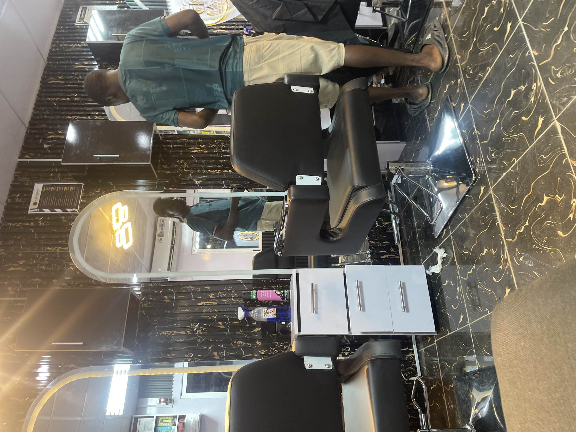 The image shows a dimly lit barbershop in Barako, Nigeria. In the foreground, a black barber chair with chrome accents is partially visible. To the left of the chair is a white trolley with two drawers, each with a silver handle. On top of the trolley sit two bottles, one blue and one pink, both with spray nozzles. The floor is tiled with black and gold marble-patterned tiles.
In the mid-ground, a man wearing a teal t-shirt and light-colored shorts stands with his back to the camera, facing a large mirror. Behind him, reflected in the mirror, is another man, presumably a barber, wearing a similar teal t-shirt and also facing away from the camera. Another barber chair is visible in the reflection. The walls are decorated with black and gold patterned wallpaper. To the left of the main mirror, a tall, narrow mirror with a yellow frame is visible, reflecting a shelf of products.
In the background, there are several black cabinets mounted on the wall, with silver handles. Above the main mirror, a neon sign in the shape of the number "88" glows brightly. Further to the right, a white air conditioning unit is mounted high on the wall. A black cape is draped over a chair on the far right. The overall atmosphere suggests a professional grooming establishment.