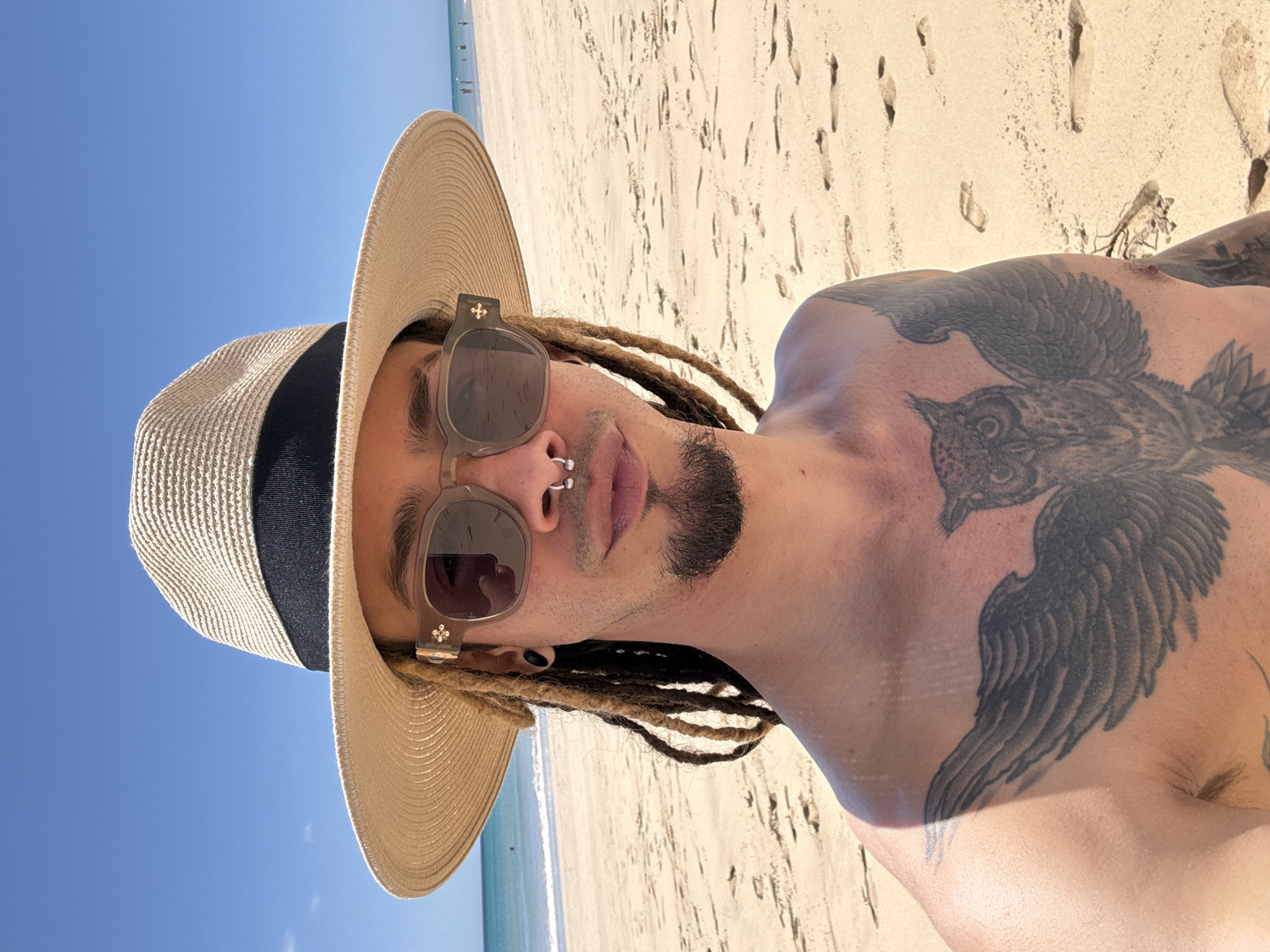 This image, rotated 90 degrees clockwise, presents a person relaxing on a bright, sunny beach in Pájara, Spain. The subject, depicted from the chest up, has light brown dreadlocks visible under a wide-brimmed, light beige straw hat with a dark blue band. They wear stylish grey-tinted sunglasses featuring a distinctive small cross emblem on the side hinges.
The person has a well-groomed goatee and mustache, a horseshoe-shaped septum piercing, and a dark stud in their visible left ear. They are shirtless, prominently displaying a large, intricate tattoo of an owl with outstretched wings that spans across their chest and left shoulder.
The background is a vibrant split of a clear, cloudless blue sky on the left and a expansive, light beige sandy beach on the right. The sand is marked with numerous footprints, suggesting activity. In the distance, the calm, turquoise ocean meets the shore, with hints of small figures (likely other beachgoers) near the waterline. The strong, direct sunlight indicates a clear and warm daytime setting, conveying a relaxed and pleasant atmosphere. No visible text is present.