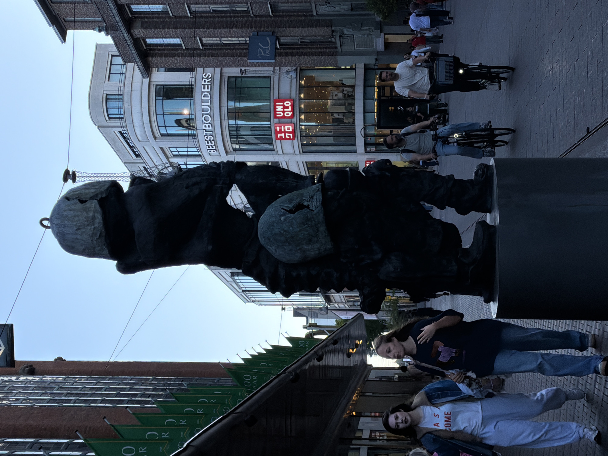 The image shows a street scene in The Hague, Netherlands, likely on a shopping street given the presence of shops and people. In the foreground, a large, dark, abstract sculpture stands on a pedestal. To the left of the sculpture, two young women walk side-by-side on a paved sidewalk. The woman on the left wears a denim jacket over a white t-shirt and light-colored sweatpants. The woman on the right wears a dark t-shirt and jeans. Behind them, a series of green banners with white text are hanging from a building. Further down the street, two men are riding bicycles. The man on the left wears a gray t-shirt and jeans, and the man on the right wears a white t-shirt and dark pants. They are riding on a brick-paved street. In the background, there are multi-story buildings. One building has prominent signage that reads "BEESTBOULDERS" and "UNI QLO" with red logos. The sky is a clear, light blue, suggesting it is daytime, possibly late afternoon or early evening due to the lighting. The overall impression is a bustling urban environment.