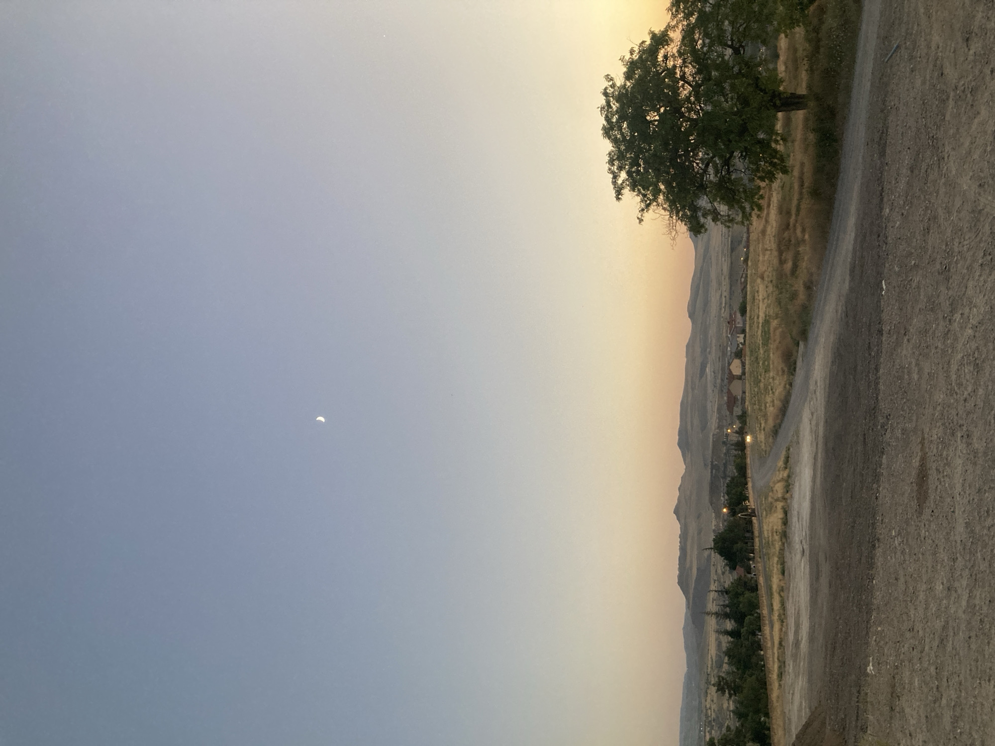 An unobstructed, eye-level, wide shot captures a rural landscape near Avanos İlçe Merkezi, Turkey, at twilight. The sky transitions from a pale blue at the top to a soft orange and yellow near the horizon. A sliver of the moon is visible in the upper left quadrant of the sky. In the middle ground, a dirt road winds through dry, golden grass. To the right, a large, leafy tree stands prominently. In the distance, a range of soft, rolling hills forms the backdrop, with small buildings and scattered trees visible along the slopes and in the valley. The foreground is dominated by a rough, dusty patch of ground. There are no people or animals present in the scene. The overall impression is one of serene, natural beauty at the close of day.