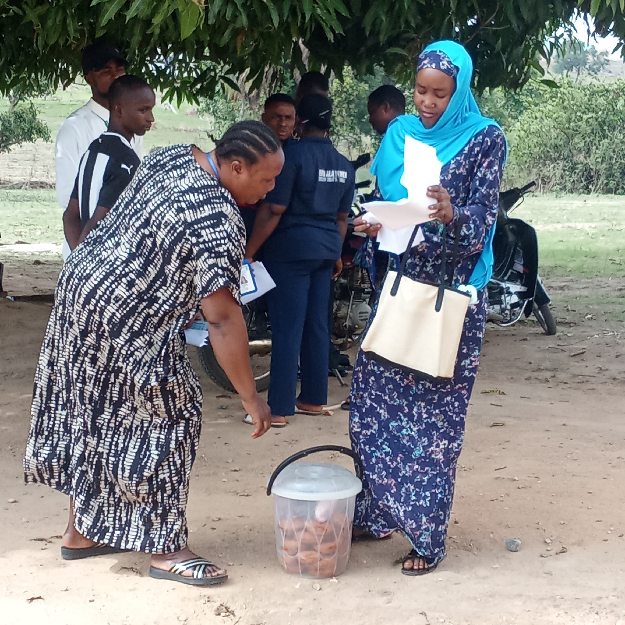 The image shows a group of people gathered outdoors in an area appearing to be in the city of Unknown, Unknown. The scene takes place on a dirt ground surface under a tree providing some shade. A woman in a blue headscarf and a patterned blue dress stands on the right side of the image, holding papers and a cream-colored bag. Beside her is a clear plastic bucket filled with what appear to be baked goods or snacks. Another woman, wearing a black and white patterned dress, is looking down towards the bucket. Several people are standing in the background, including one man in a white shirt and another in a black and white soccer jersey. A motorcycle is parked behind the group.
