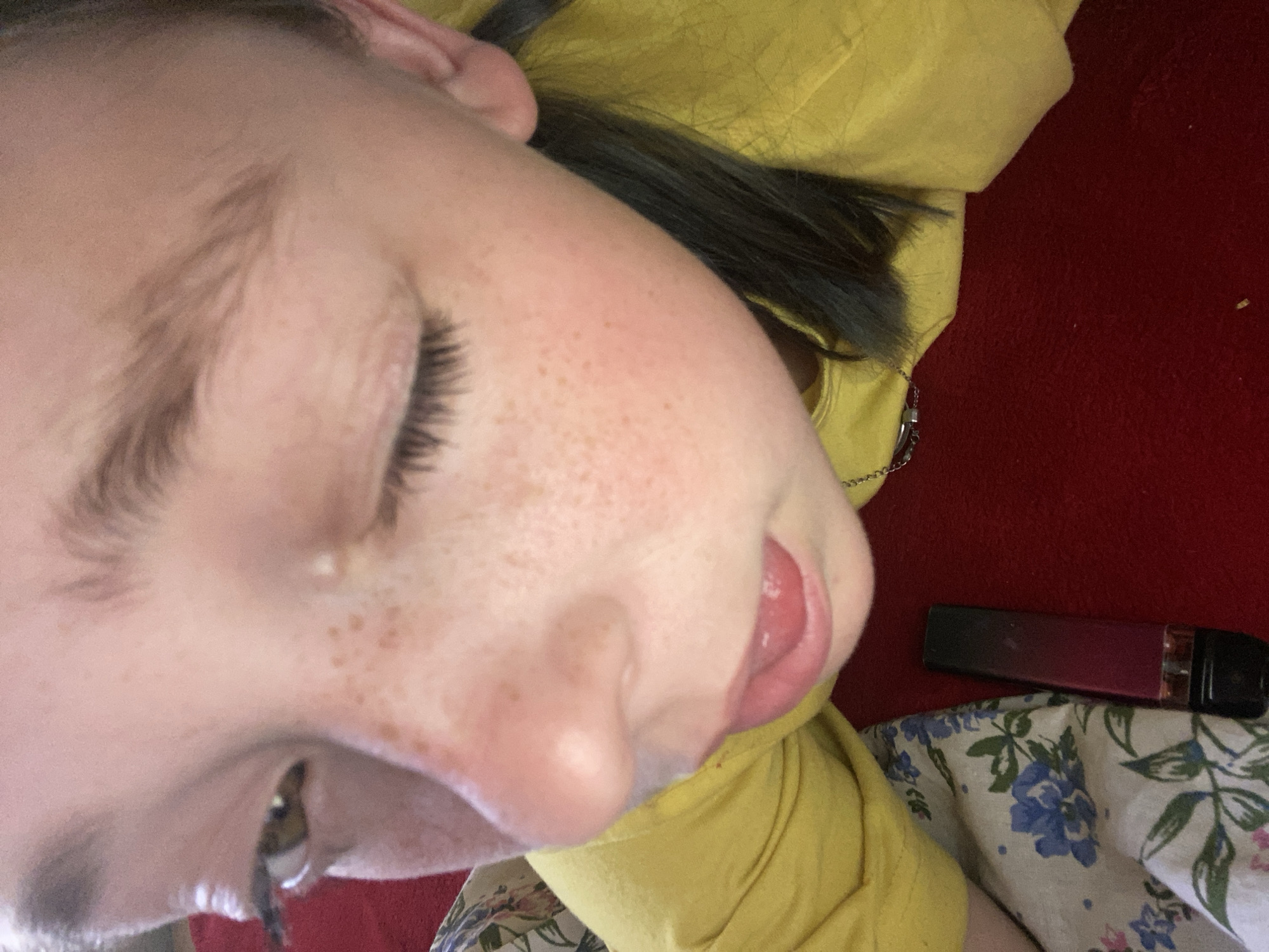 The image shows a close-up, low-angle shot of a person's face, with their head tilted. They have fair skin with freckles across their nose and cheeks. Their eyes are closed, and their eyelashes are dark and prominent. The person's tongue is slightly extended, and their lips are a light pink color. Dark hair is visible, some of which appears to be pulled back. The person is wearing a yellow garment, possibly a hoodie or sweater.
In the background, a red surface is visible, which could be a rug or carpet. To the right, a dark red, cylindrical object with a black cap is lying on a white fabric with a floral pattern featuring blue flowers and green leaves. The lighting suggests an indoor setting, possibly with natural light. No specific time of day or weather conditions can be determined. There is no visible text in the image. The setting is identified as Nova Praha, Ukraine. The activity appears to be a moment of rest or relaxation.