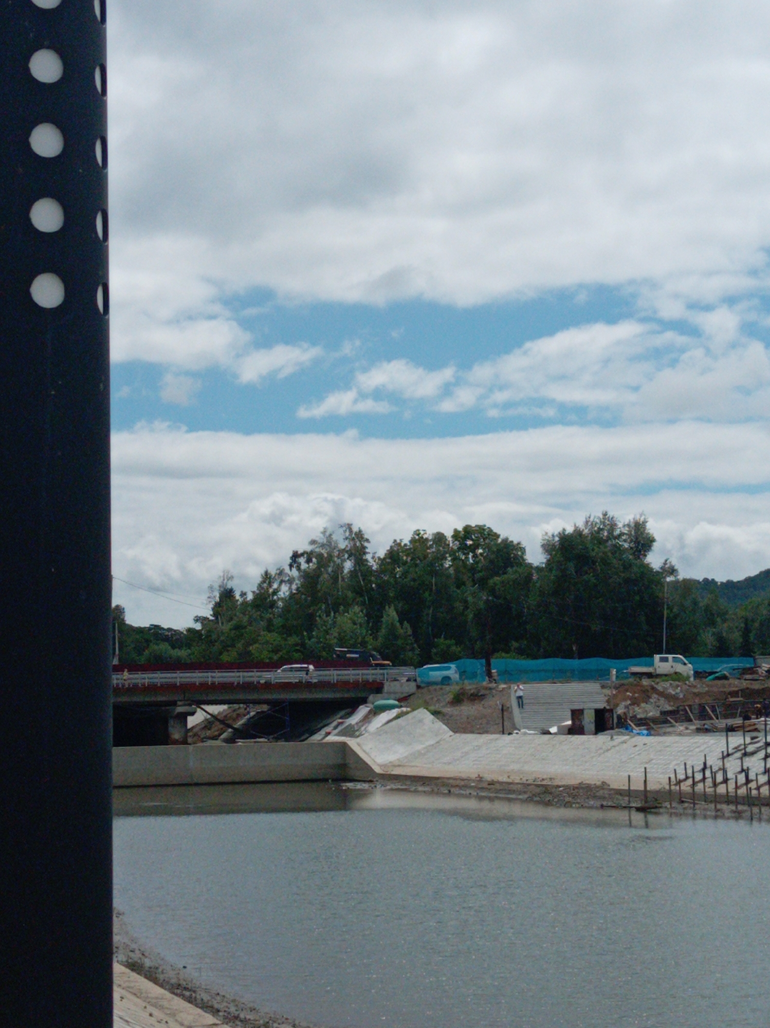 The image shows a construction site along a body of water, with a bridge in the background. A large, dark, cylindrical object with white circular cutouts is in the foreground on the left side of the frame, obscuring a portion of the scene. The body of water appears to be a river or harbor. On the far bank, there is a concrete embankment with some construction materials and equipment visible, including a white van and what appears to be a small building or shelter. Further back, a red bridge spans the water, with several vehicles on it. Dense green trees line the horizon behind the bridge, with a hillside visible in the distance. The sky is cloudy with patches of blue. This scene is consistent with areas of Artyom, Russia, that are undergoing infrastructure development or are situated near waterways.