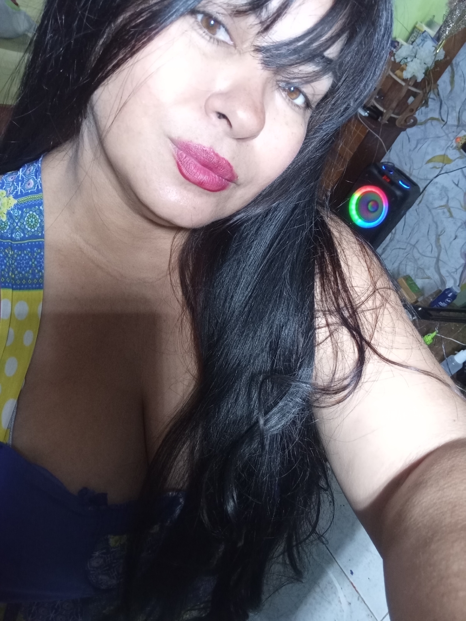 The image is a close-up, chest-up selfie of a woman with long, dark hair that frames her face and falls over her shoulders. She has brown eyes and is looking directly into the camera with her lips slightly pursed, wearing pink lipstick. Her skin tone appears warm.
She is wearing a garment with a blue, yellow, and white patterned design, possibly a dress or top, which is visible on her left side. Underneath, a solid blue or purple garment is seen, revealing cleavage.
The background depicts an indoor setting, which appears somewhat cluttered. To the right of the woman, a black portable speaker with circular, colorful LED lights displaying a rainbow effect is clearly visible. Other indistinct objects, potentially on a surface or shelf, along with some wires, are also present in the dimly lit background. The lighting primarily illuminates the woman's face. No legible text is visible in the image.