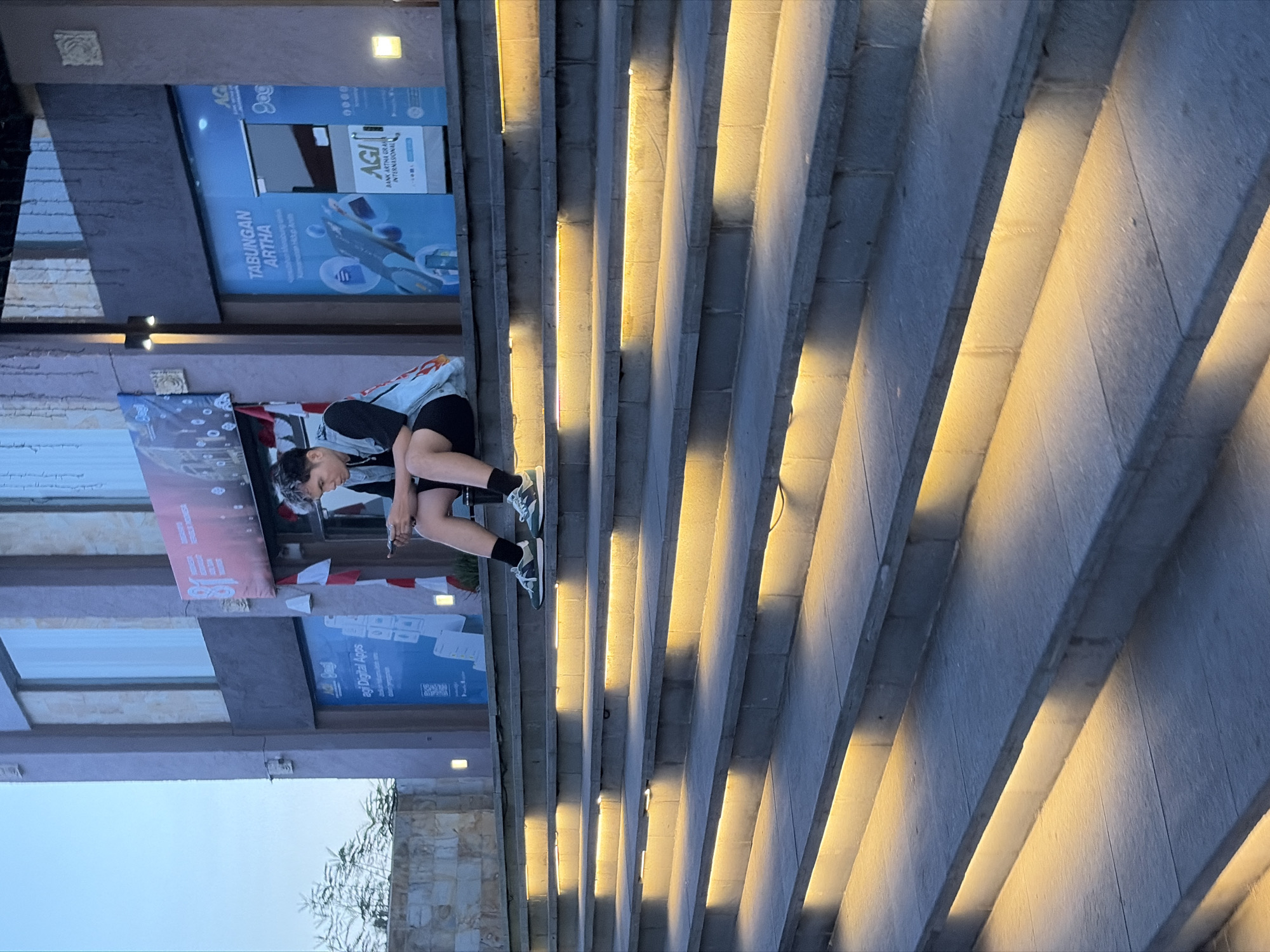 A person is sitting on a wide, illuminated staircase, looking down at their phone. The staircase is made of stone and has integrated yellow lighting along the edge of each step. Behind the staircase is a building with a modern facade, featuring large windows and signage. One sign, partially visible, advertises "AGI BANK ARTHA INTERNATIONAL" and another appears to be a promotional banner. The person is wearing a dark t-shirt, a light-colored vest with a fiery graphic, dark shorts, black socks, and athletic shoes. They have dark hair with lighter streaks and appear to be in their late teens or early twenties. The overall scene suggests an outdoor public space, possibly a commercial or entertainment area in Kuta, Indonesia, given the signage. The lighting indicates it is either dusk or evening.