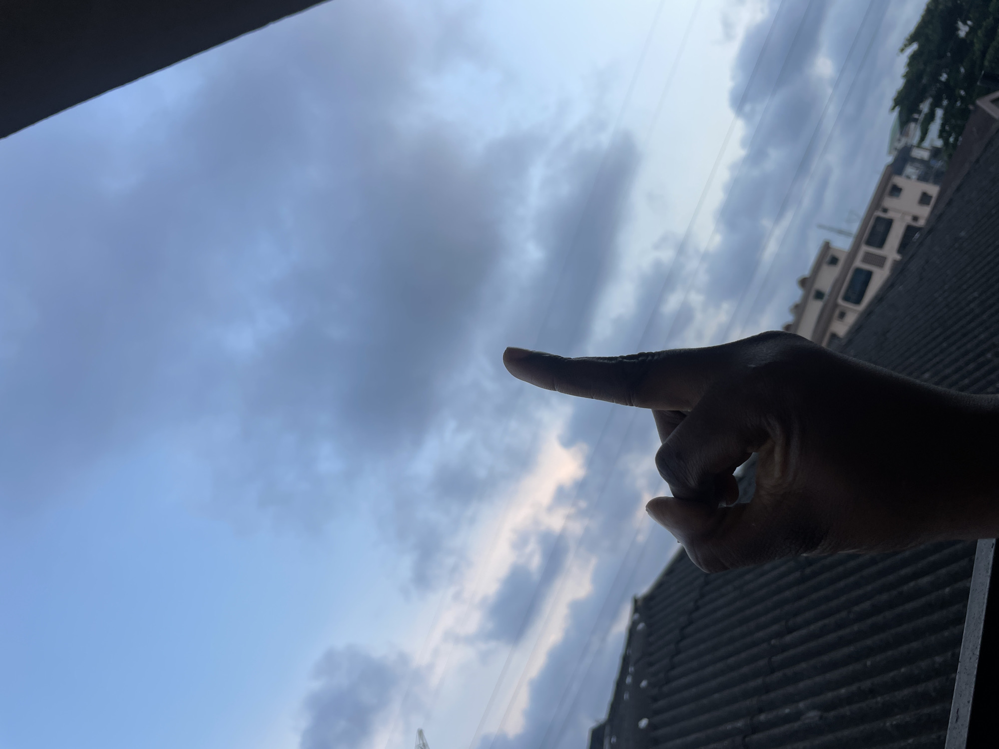 The image shows a person's hand with the index finger extended upwards, silhouetted against a cloudy sky. In the background, there is a sloped, dark-colored roof of a building, and further beyond, several multi-story buildings and trees are visible. The sky is a pale blue with scattered, darker clouds, suggesting it might be dawn or dusk. The scene appears to be outdoors, likely in an urban or semi-urban environment in Animashawun, Nigeria, given the architectural style of the buildings and the general atmosphere. The hand is the primary subject in the foreground, with the finger pointing towards the sky. The lighting is dim, indicating low natural light conditions.