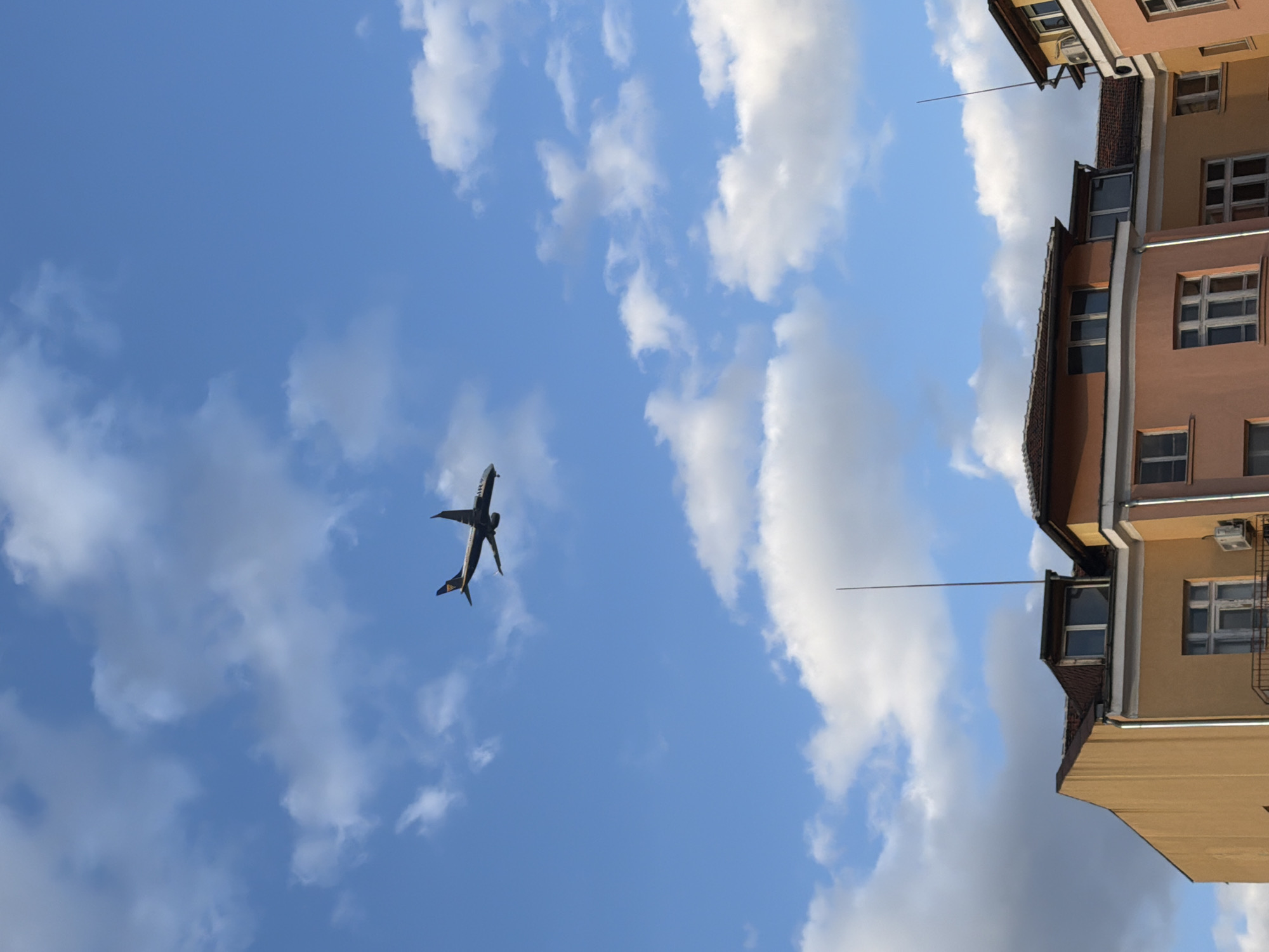 This image captures an urban scene in Sofia, Bulgaria, under a partly cloudy blue sky. A commercial airliner, dark in color with a yellow logo visible on its tail fin, is in mid-flight in the upper left quadrant, oriented as if ascending or descending, with its underside exposed. The lower third of the frame reveals the upper sections of multiple multi-story residential buildings. The leftmost building exhibits an ochre-yellow facade. Adjacent to it is a peach-colored building featuring white trim around its multi-paned windows, a dark-railed balcony, and an external air conditioning unit mounted on its facade. To the right, another section of a yellow building is partially visible, followed by a further peach-colored structure. All visible buildings are topped with terracotta-tiled roofs. Two slender, dark-colored lightning rods extend vertically from the rooftops of the central and rightmost buildings towards the sky. No individuals are visible in the scene. The lighting suggests daytime conditions.