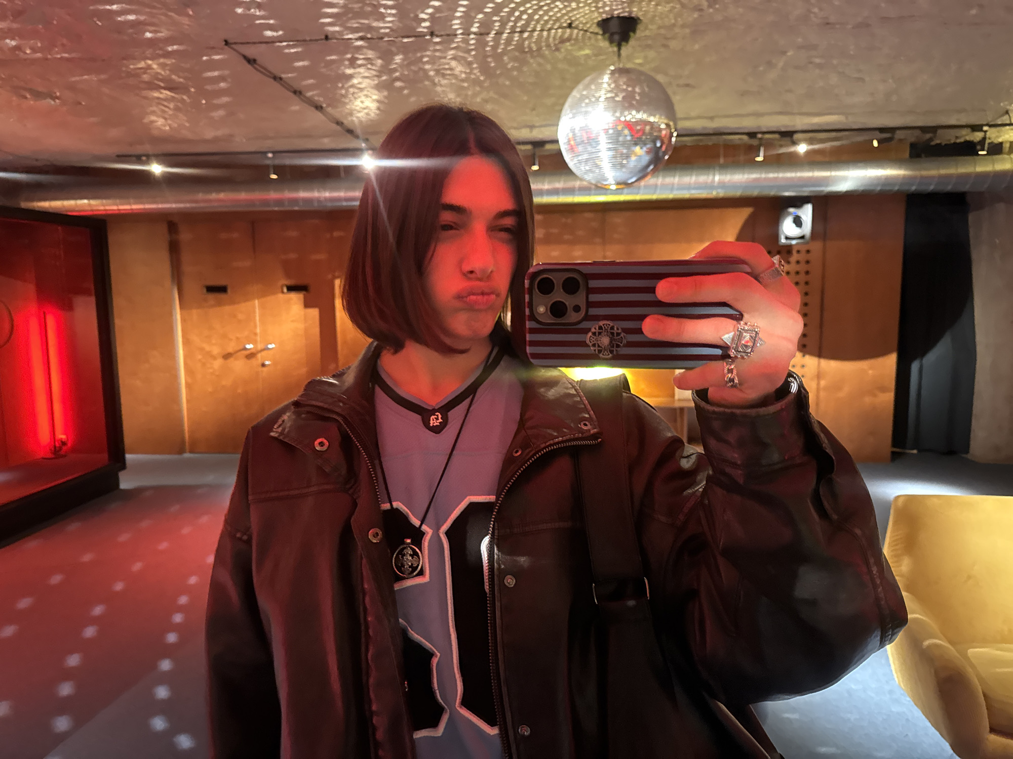 A person with short brown hair and fair skin is taking a selfie indoors. They are wearing a brown leather jacket over a blue and white jersey-style shirt, with a black cord necklace bearing a pendant. The person is holding a smartphone with a striped case, adorned with a decorative emblem. They have multiple rings on their fingers.
The background features a room with a disco ball hanging from the ceiling, which reflects patterns of light onto the textured ceiling. The walls appear to be paneled with wood, and a metal duct is visible. In the left background is a red-lit display case. To the right is a yellow chair. The setting is identified as Yerevan, Armenia.