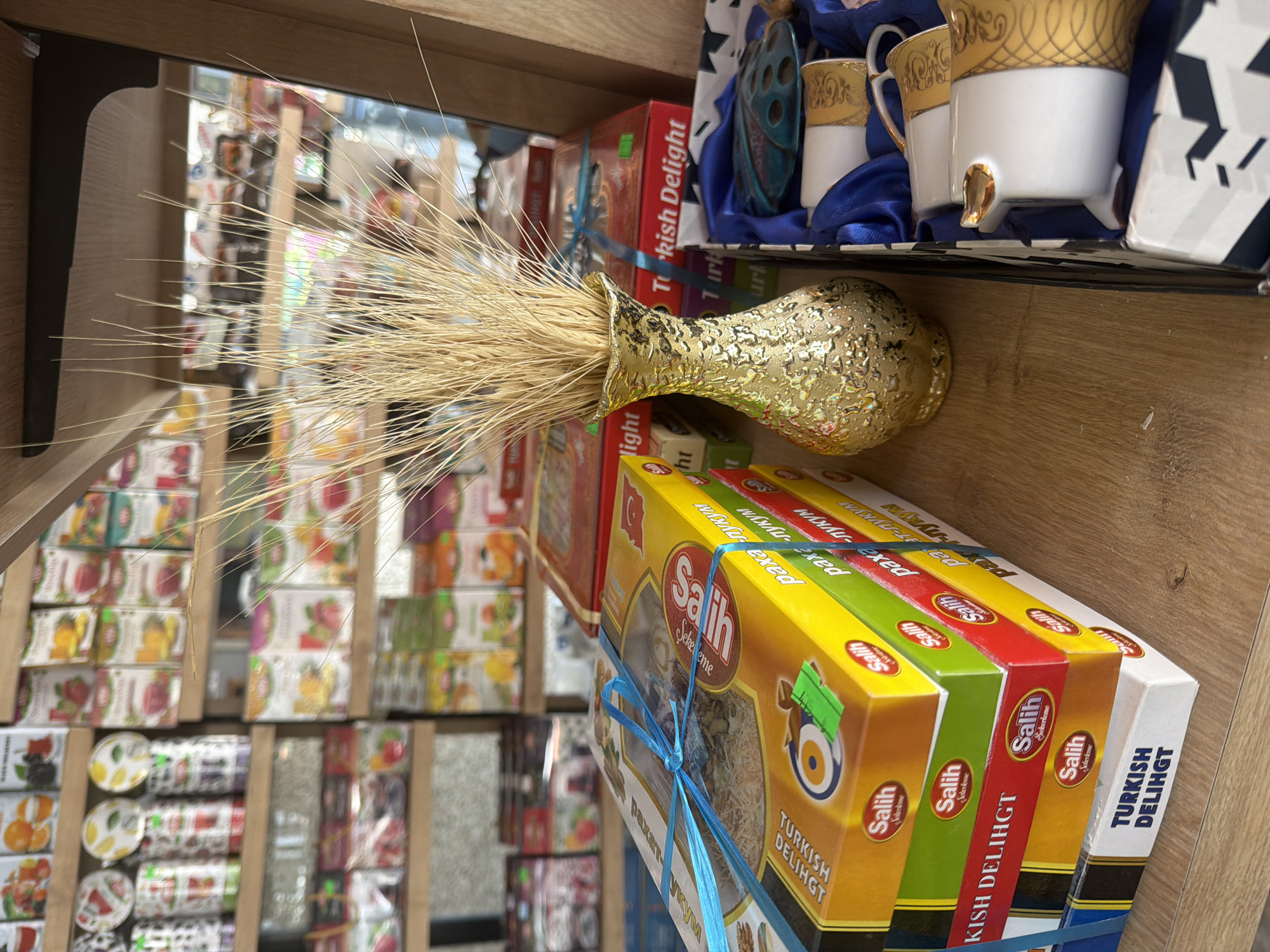 In a retail setting, likely a shop in Marmaris, Turkey, a wooden shelf displays an assortment of packaged goods, primarily boxes of Turkish Delight. A stack of these boxes, some tied with blue ribbon, dominates the foreground, with visible branding including "Salih" and "Turkish Delight." To the right of the boxes, a gold, textured vase holds a bundle of dried wheat stalks. Next to the vase, a gift set containing white ceramic cups with gold embellishments is presented in a box lined with blue satin. The background shows shelves filled with more packaged items, some appearing to be tea boxes with colorful fruit imagery. The lighting suggests an indoor environment, possibly a market or a specialty store.